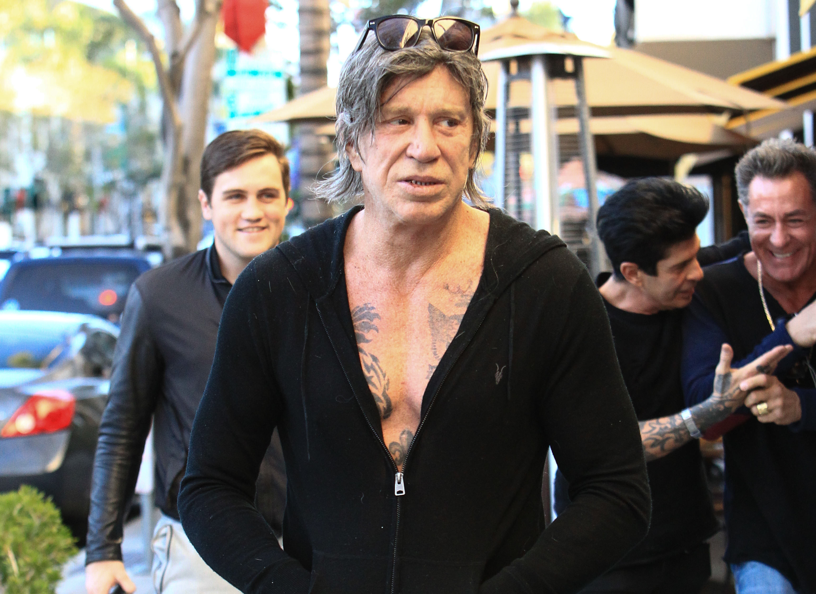 Mickey Rourke is seen in Los Angeles sporting a casual black zip-up hoodie left open to reveal his chest tattoos. With sunglasses perched on his head and a serious gaze, he exudes rugged cool while strolling through the city.