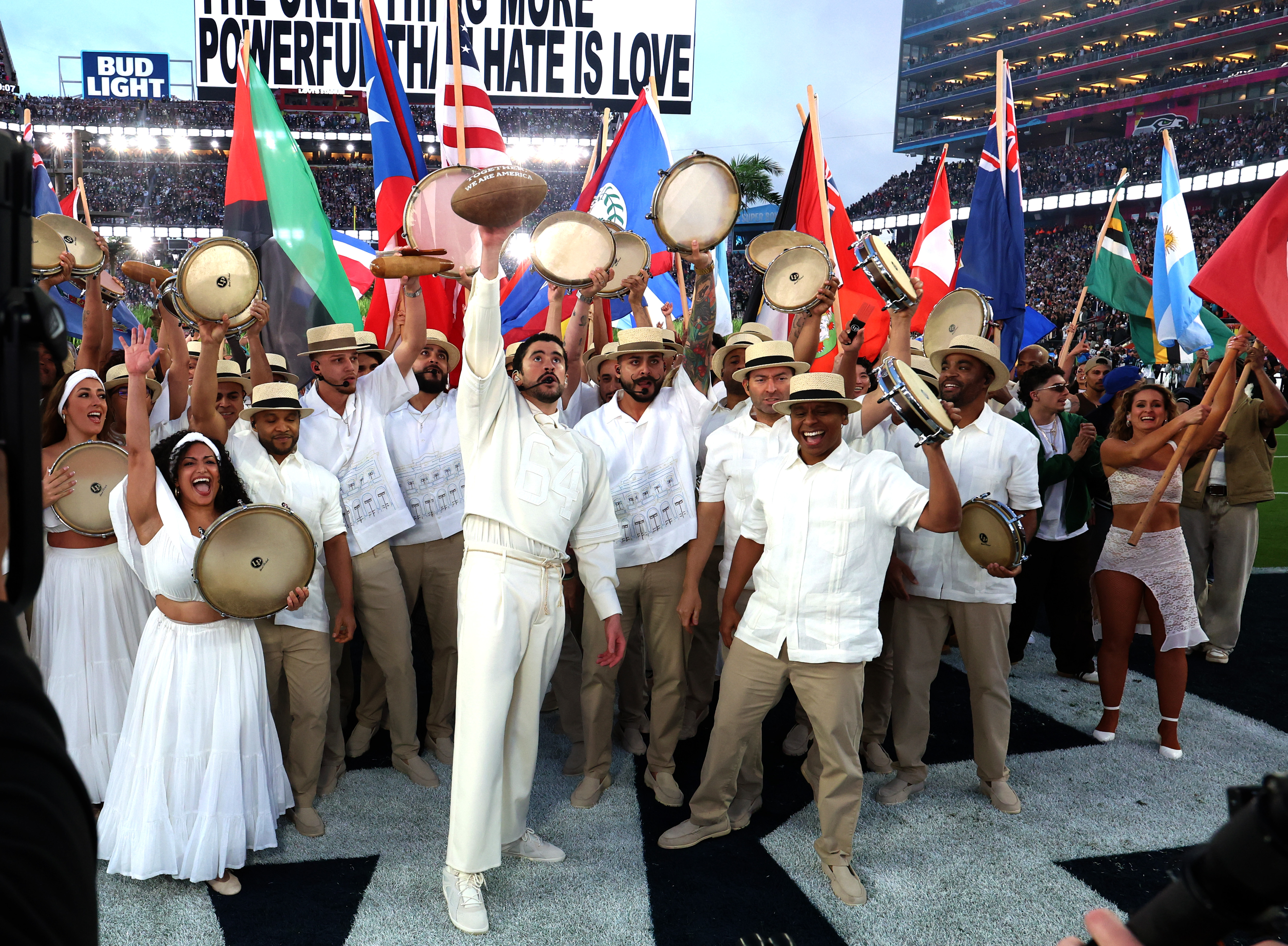 Bad Bunny performs with tambourine-holding dancers and flag bearers, raising his hand while surrounded by performers celebrating on the field during the halftime show. | Source: Getty Images