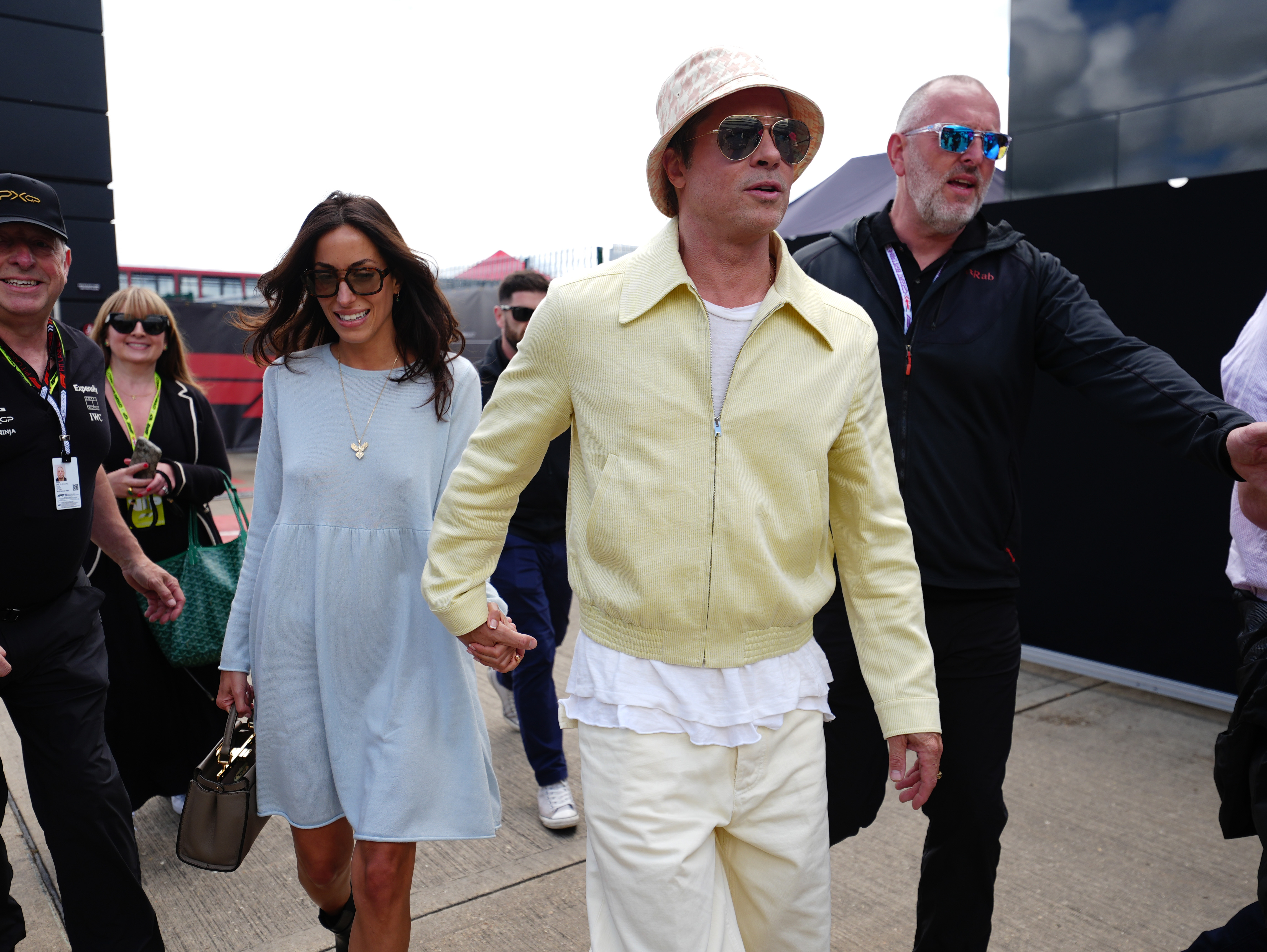 Brad Pitt and his partner Ines de Ramon walked hand-in-hand as they arrived at Silverstone Circuit in Northamptonshire on July 7, 2024. Pitt sported a light yellow zip-up jacket over a white ruffled hem, cream trousers, aviator sunglasses, and a houndstooth-patterned bucket hat, while de Ramon walked alongside him in a pastel blue long-sleeve shift dress, oversized dark sunglasses, and a pendant necklace. | Source: Getty Images