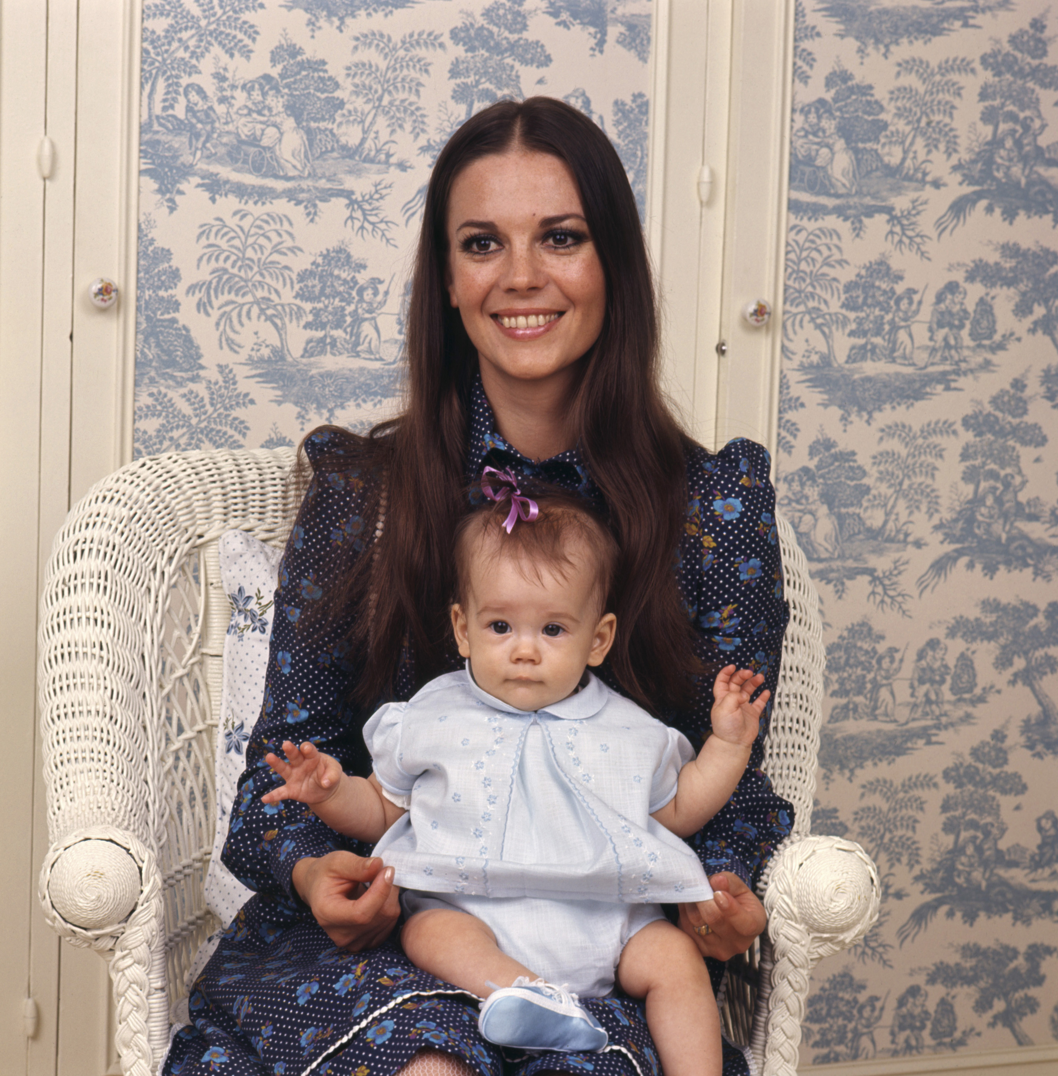 Natalie Wood sits in a white wicker chair, holding baby Natasha Gregson Wagner on her lap. Dressed in a dark, patterned dress with blue and white floral details, Natalie smiles softly at the camera. Baby Natasha wears a pale blue outfit with embroidered flowers, a purple bow in her hair, and matching blue booties.