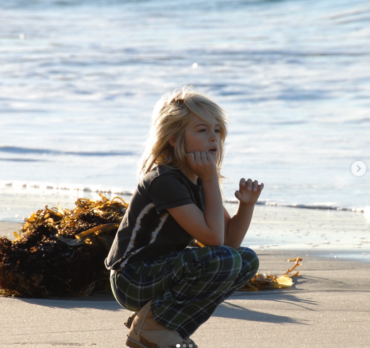 Crouched near a mound of seaweed along the shore, a young, blonde-haired Presley Gerber rests his chin on his hand while looking out toward the tide in a nostalgic beach snapshot posted on June 25, 2025. | Source: Instagram/presleygerber