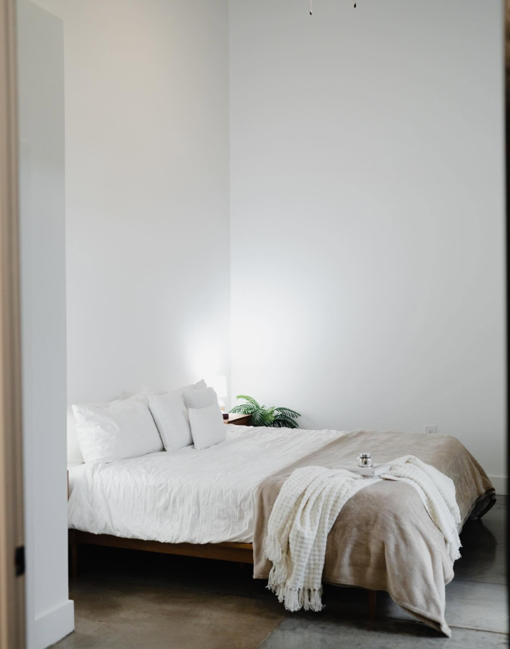 A serene bedroom offers a softer side of the loft, with a low wooden bed, neutral bedding and a small touch of greenery against the otherwise minimalist backdrop. | Source: Facebook/Judson Mills Loft