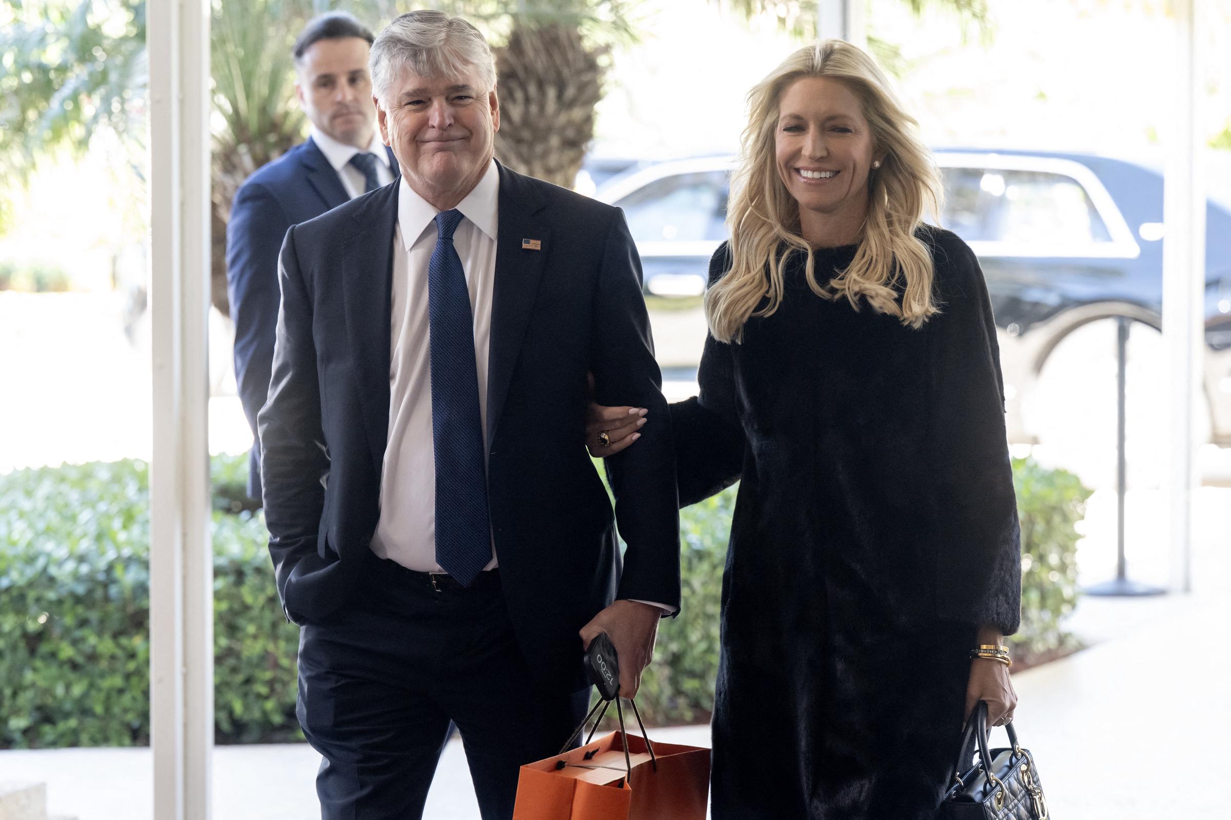 Ainsley Earhardt smiles as she links arms with fiancé Sean Hannity while arriving to attend the wedding of Dan Scavino and Erin Elmore at Mar-a-Lago in Palm Beach, Florida, on February 1, 2026, dressed in an elegant textured black midi dress paired with a black quilted leather Lady Dior handbag and styled with soft blonde waves. | Source: Getty Images