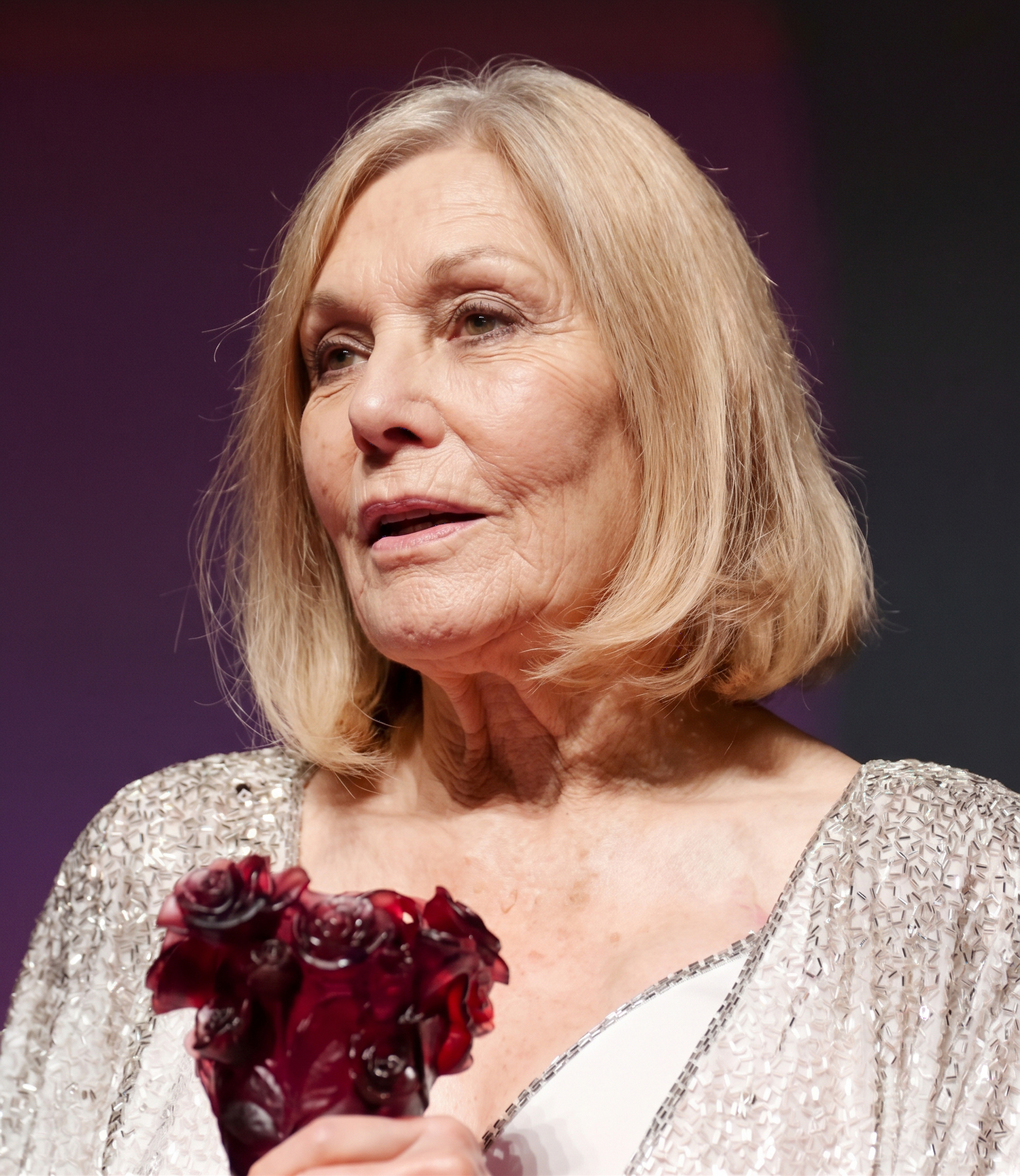 Kim Novak turns slightly to the side in a shimmering silver jacket and white dress, holding a dark red floral award against a muted purple backdrop. | Source: Gemini