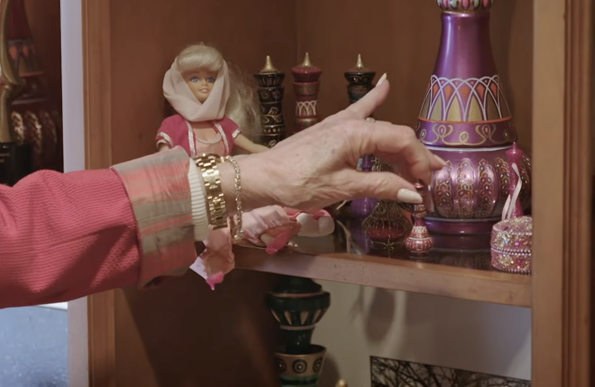 A close-up of Barbara Eden's collection shows her reaching toward a miniature decorative bottle beside a blonde doll dressed in pink and an elaborate purple-and-gold genie bottle. | Source: YouTube/People