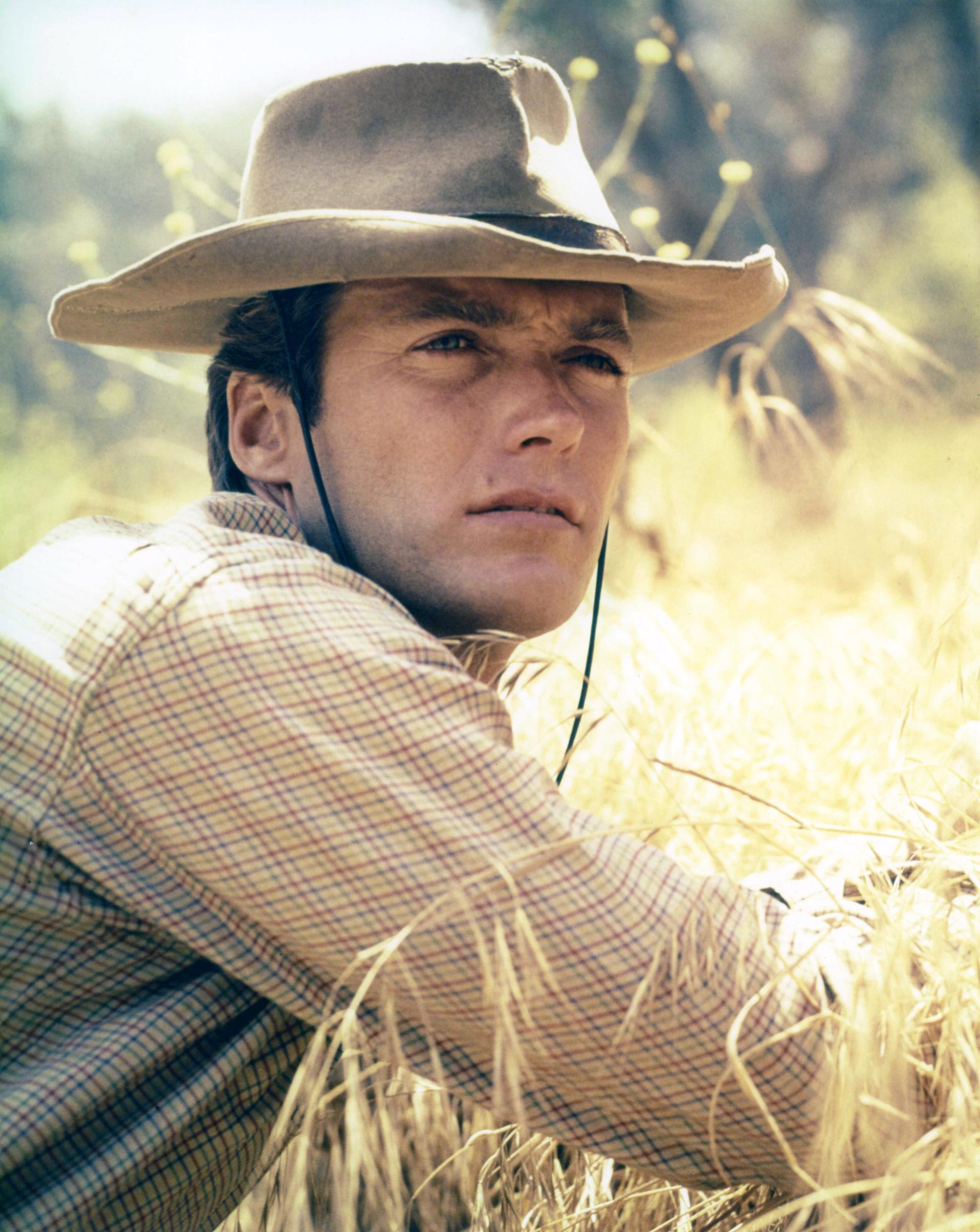 Clint Eastwood embraced his Western look on the set of the TV series "Rawhide" circa 1959, wearing a wide-brimmed cowboy hat and checked shirt while resting amid tall, sunlit grass. Then in his late 20s, the rising Hollywood actor stared intently into the distance, with his piercing eyes, strong brow, prominent cheekbones, and chiseled jawline standing out.