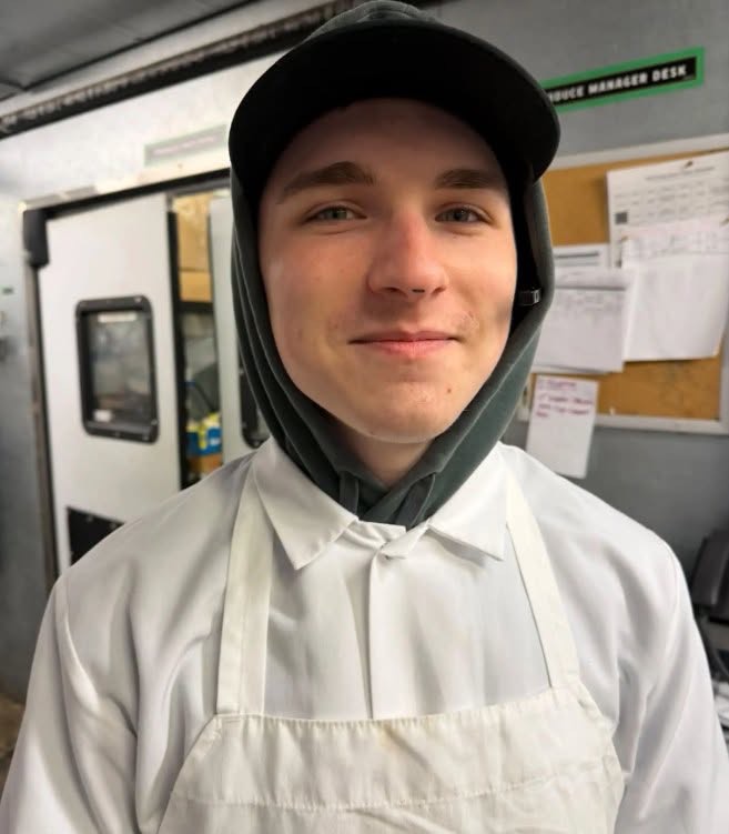 Maclaine Schmidt is pictured wearing a white apron, hoodie, and baseball cap in what appears to be a workplace — a glimpse into the young man's everyday life. | Source: Facebook / Carlos Moreno Reporta.