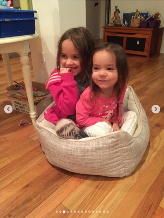 Mabel and Fiona are pictured together as young sisters, squeezed into a small upholstered chair and smiling for the camera. The candid childhood moment captures the easy closeness between them, with Mabel sitting just behind Fiona as they share an ordinary day at home.