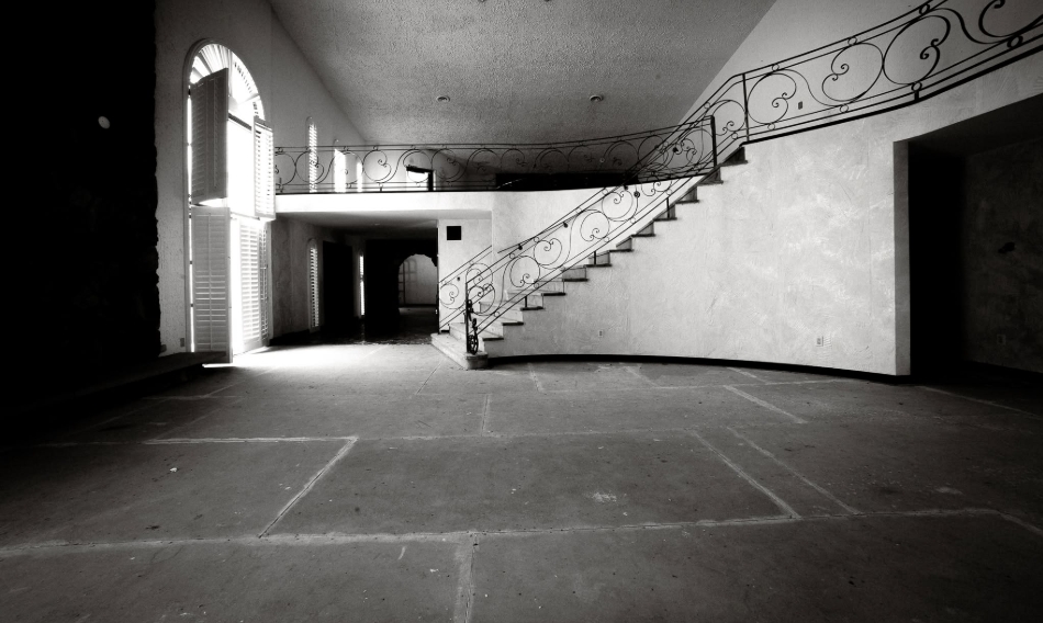A sweeping staircase curves toward the upper floor of Mike Tyson's former mansion, bordered by decorative wrought-iron railings that continue around the overlooking balcony. Tall arched windows illuminate the otherwise bare interior, where removed furnishings left the home's architectural details exposed. The scene captured how the once-opulent property appeared after years without the luxurious possessions associated with Tyson's time there. | Source: Facebook/Johnny Joo