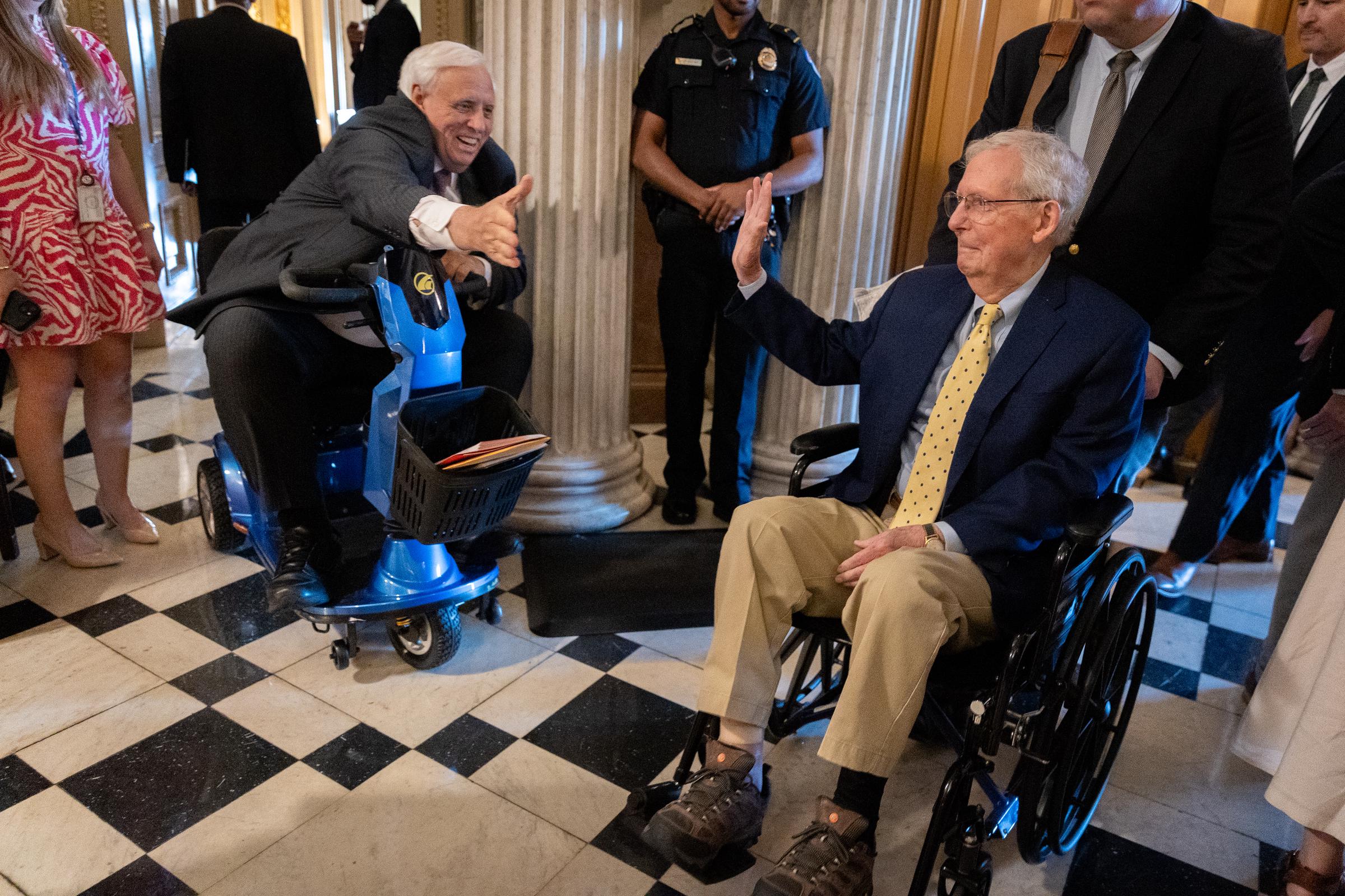 Senator Mitch McConnell waves while being pushed through the Capitol in a wheelchair, as Sen. Jim Justice gives him a thumbs-up from a mobility scooter. | Source: Getty Images