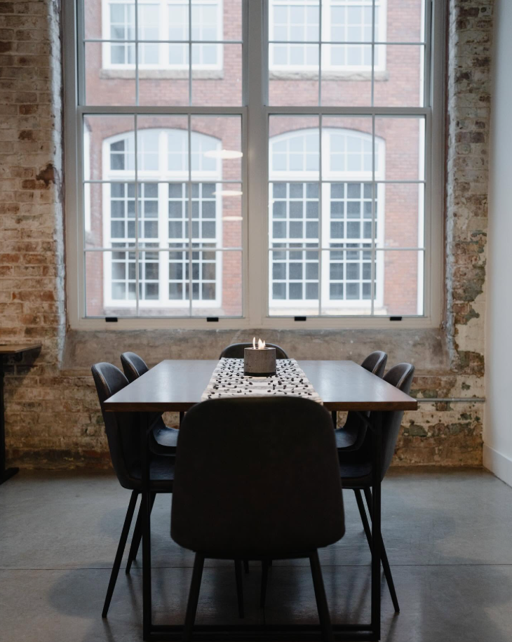 A dining area inside a Judson Mill Lofts apartment features a dark wood table, black chairs and oversized windows framed by exposed brick. | Source: Facebook/Judson Mills Loft