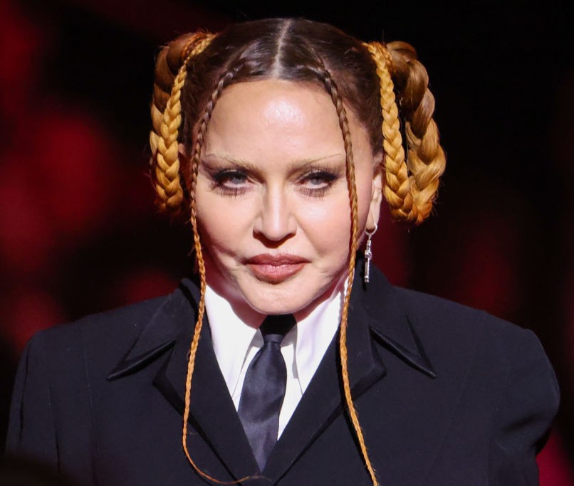 Madonna faces the camera in a black tailored suit, white shirt and black tie, with her hair styled in elaborate braided loops and long braids framing her face. | Source: Chat GPT