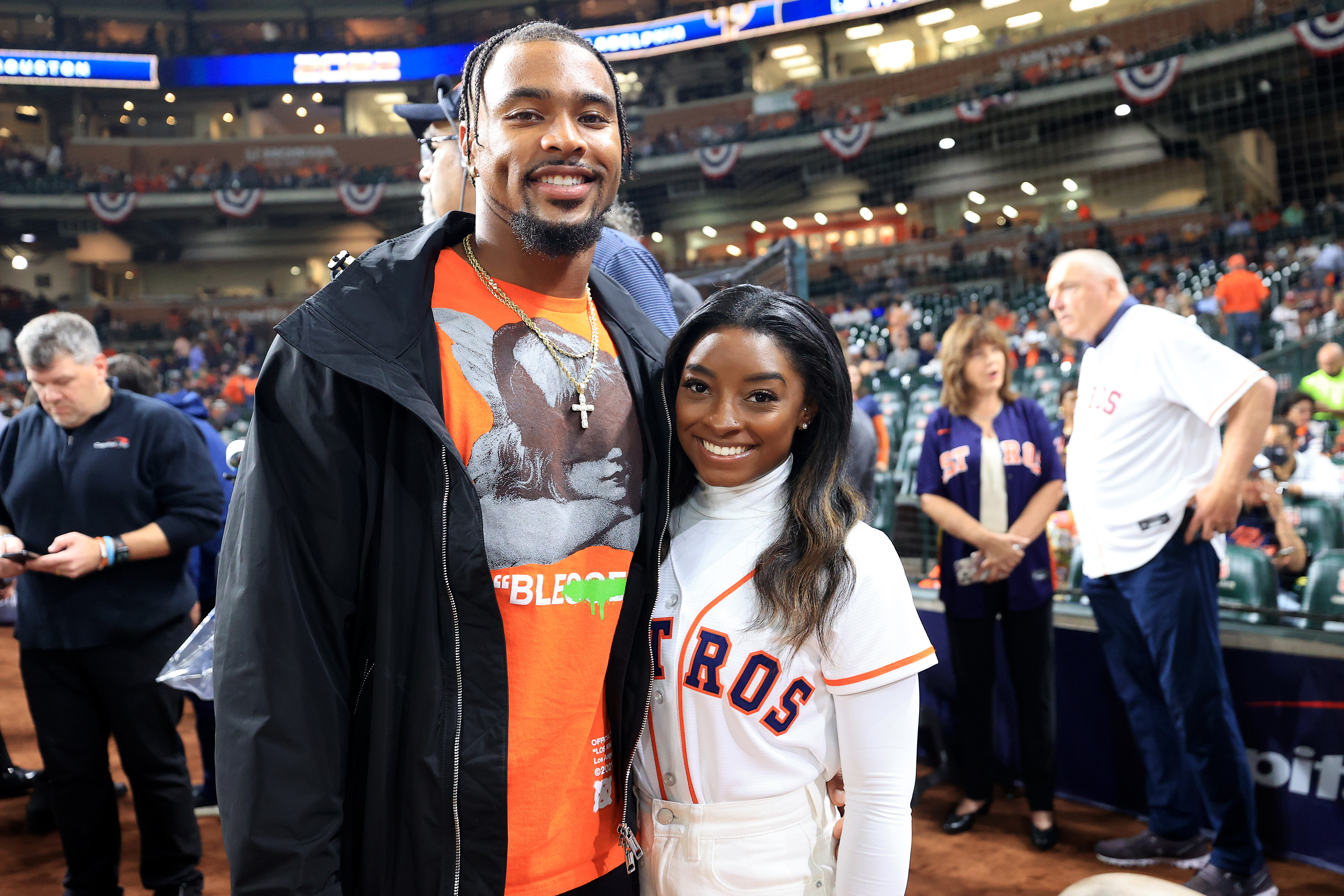 Simone Biles and Jonathan Owens smile for a photo on the field ahead of Game One of the 2022 World Series between the Philadelphia Phillies and the Houston Astros at Minute Maid Park in Houston, Texas, on October 28, 2022. Biles wears a white Astros jersey over a long-sleeve turtleneck, while Owens pairs an orange graphic tee with layered chains and a black jacket.