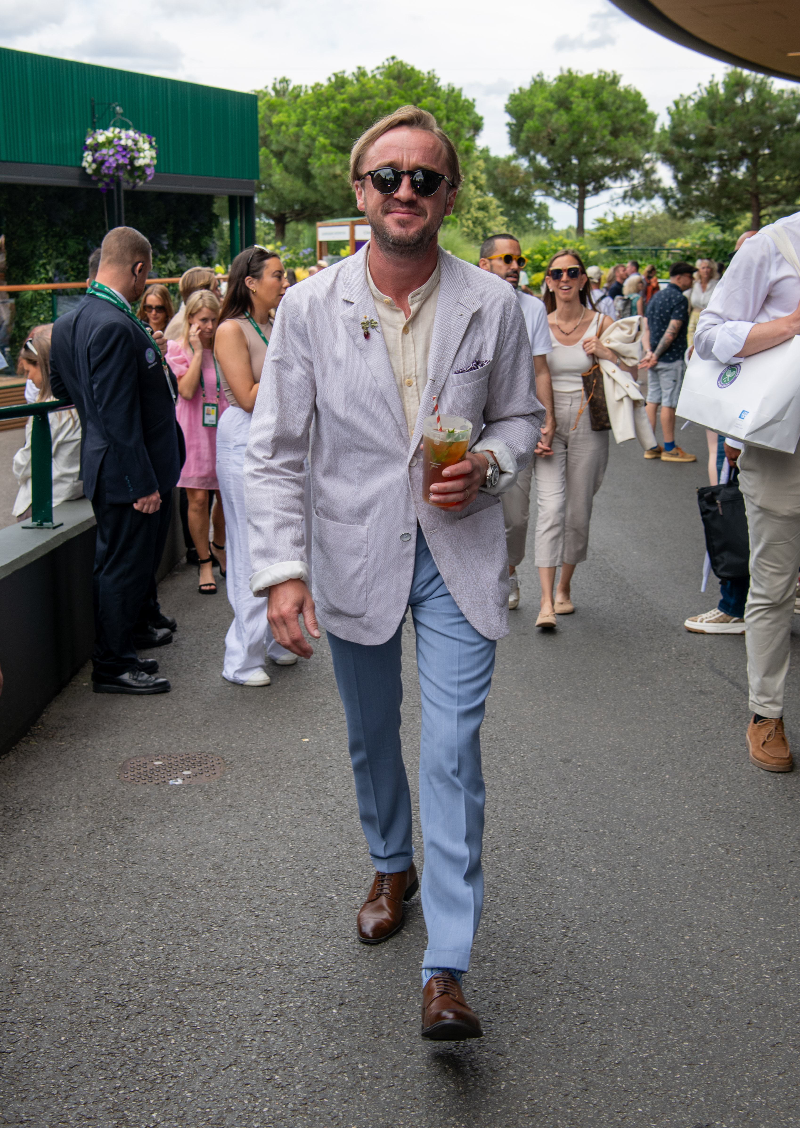 Tom Felton strolls through the Wimbledon crowds with a chilled drink in hand, blending relaxed elegance with summer charm. His light blazer, pastel trousers, and easy confidence capture the spirit of the iconic "Pimm's on the Hill" tradition.