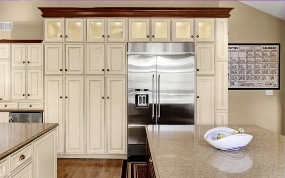 Cream-colored cabinets with raised-panel doors and dark wood trim surround a stainless-steel refrigerator in the spacious kitchen. Granite countertops and a bowl on the island add to the view of the home's traditional kitchen design. | Source: YouTube/Without A Crystal Ball