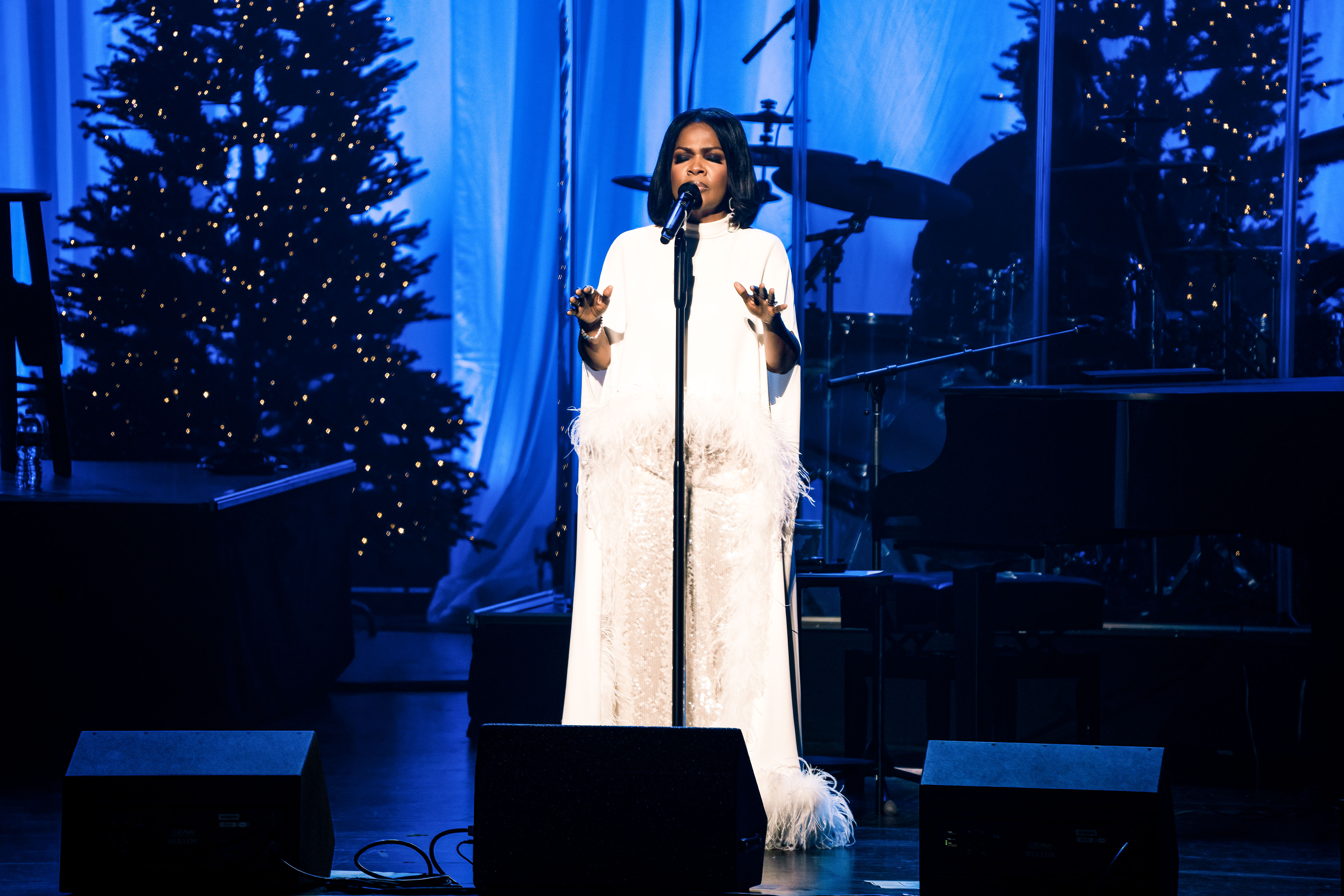 A wide shot shows CeCe Winans standing center stage with her hands lifted beside a microphone beneath blue stage lights and decorated Christmas trees as she performs before the crowd.
