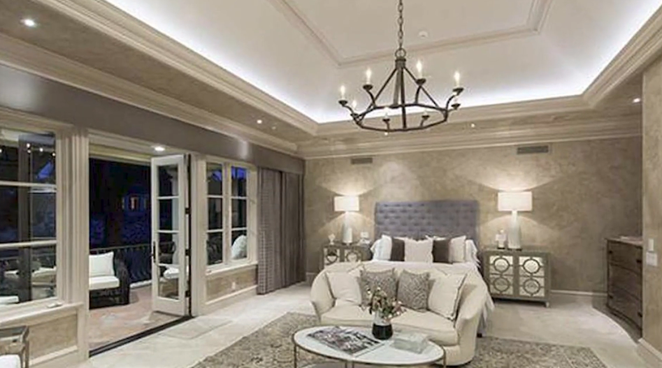 The sprawling primary bedroom suite showcases an illuminated tray ceiling anchored by a rustic wagon-wheel chandelier, paired with a tufted headboard, mirrored nightstands, an intimate sitting area, and French doors opening onto a private balustraded balcony overlooking the grounds. | Source: YouTube/Taste of Country