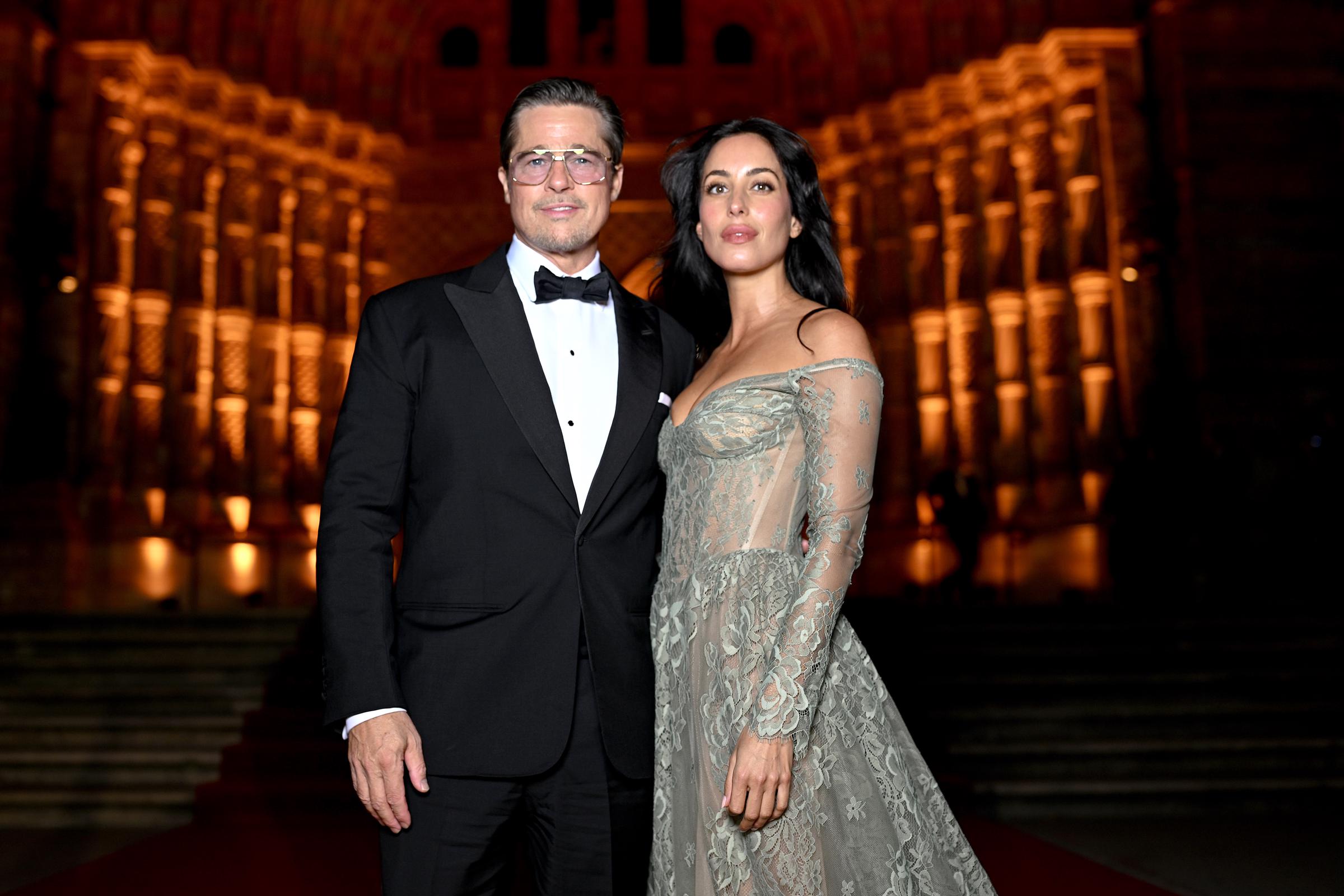 Brad Pitt and Ines de Ramon pose together on the illuminated staircase at the 2026 Albies in London, with Brad in a black tuxedo and tinted glasses and Ines wearing a sheer, off-the-shoulder lace gown with intricate floral embroidery, a fitted corset, and a flowing train. The gown's delicate detailing and translucent sleeves stand out against the venue's warm golden lighting. | Source: Getty Images
