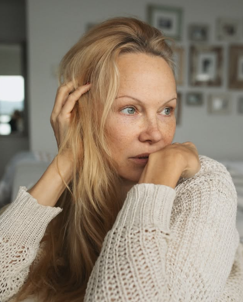 Pamela Anderson rests her chin in her hands in a contemplative, bare-faced portrait, dressed in a cozy cream open-knit sweater. Running a hand gently through her loose blonde hair against a softly blurred gallery wall of family photos, she showcases the quiet confidence and authentic beauty defining her chapter at Arcady. She paired the intimate close-up with the thoughtful reminder, "As long as we are learning — we are on the right path… 🤍✨". | Source: Instagram/pamelaanderson