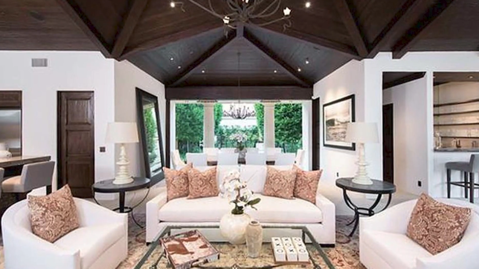 The outdoor entertaining pavilion features an exposed dark-wood beamed ceiling with a central chandelier, looking out past white columns and landscaped hedges, furnished with crisp ivory seating, patterned coral throw pillows, and an open bar pass-through. | Source: YouTube/Taste of Country