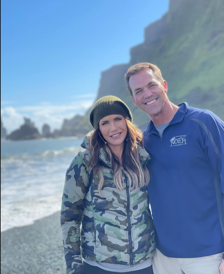 Wrapped in layers by a misty coastline, Kristi Noem and Bryon Noem lean in close — wind, waves, and warmth all in one frame. | Source: Facebook/KristiNoem