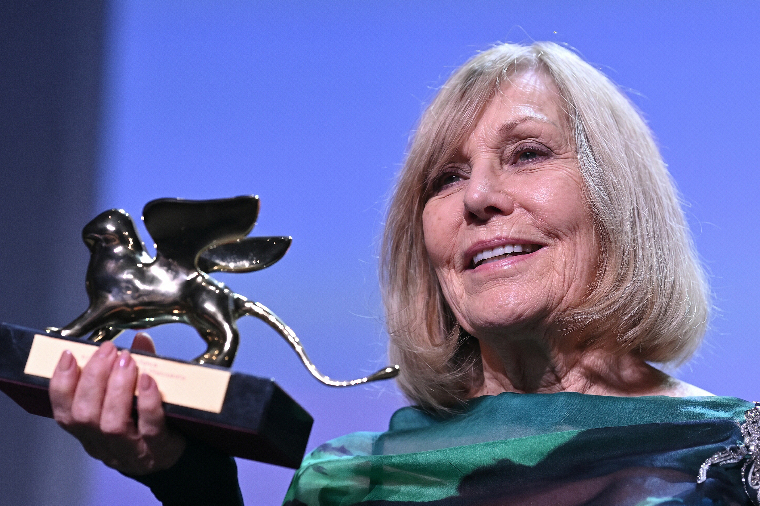 Kim Novak holds up a Golden Lion trophy and smiles in her green-and-black outfit, with the award catching the stage lights against a blue-purple background. | Source: Gemini