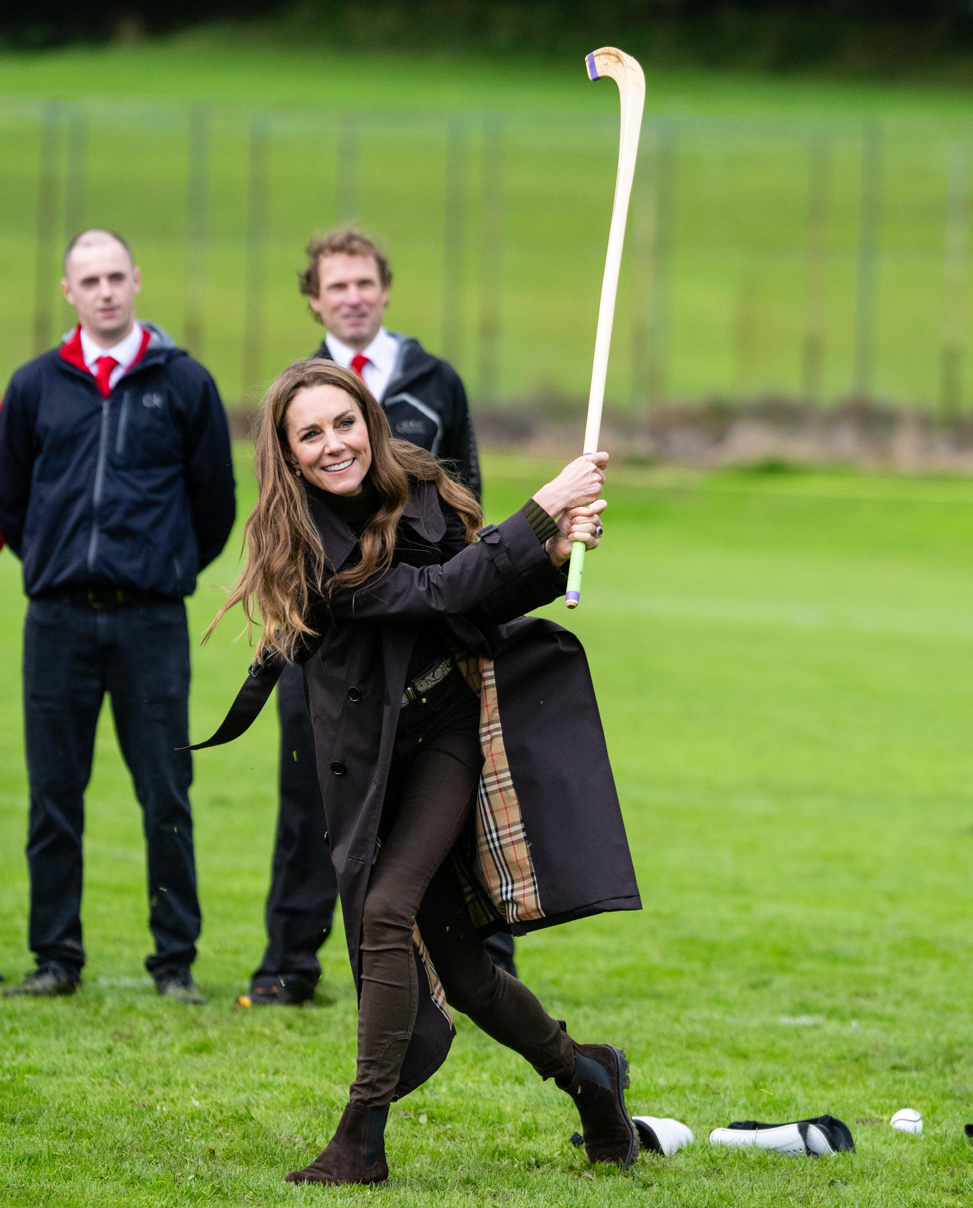 Catherine, Princess of Wales, holds the shinty stick high after her swing, smiling toward the action as players and club members look on from the pitch. | Source: Getty Images