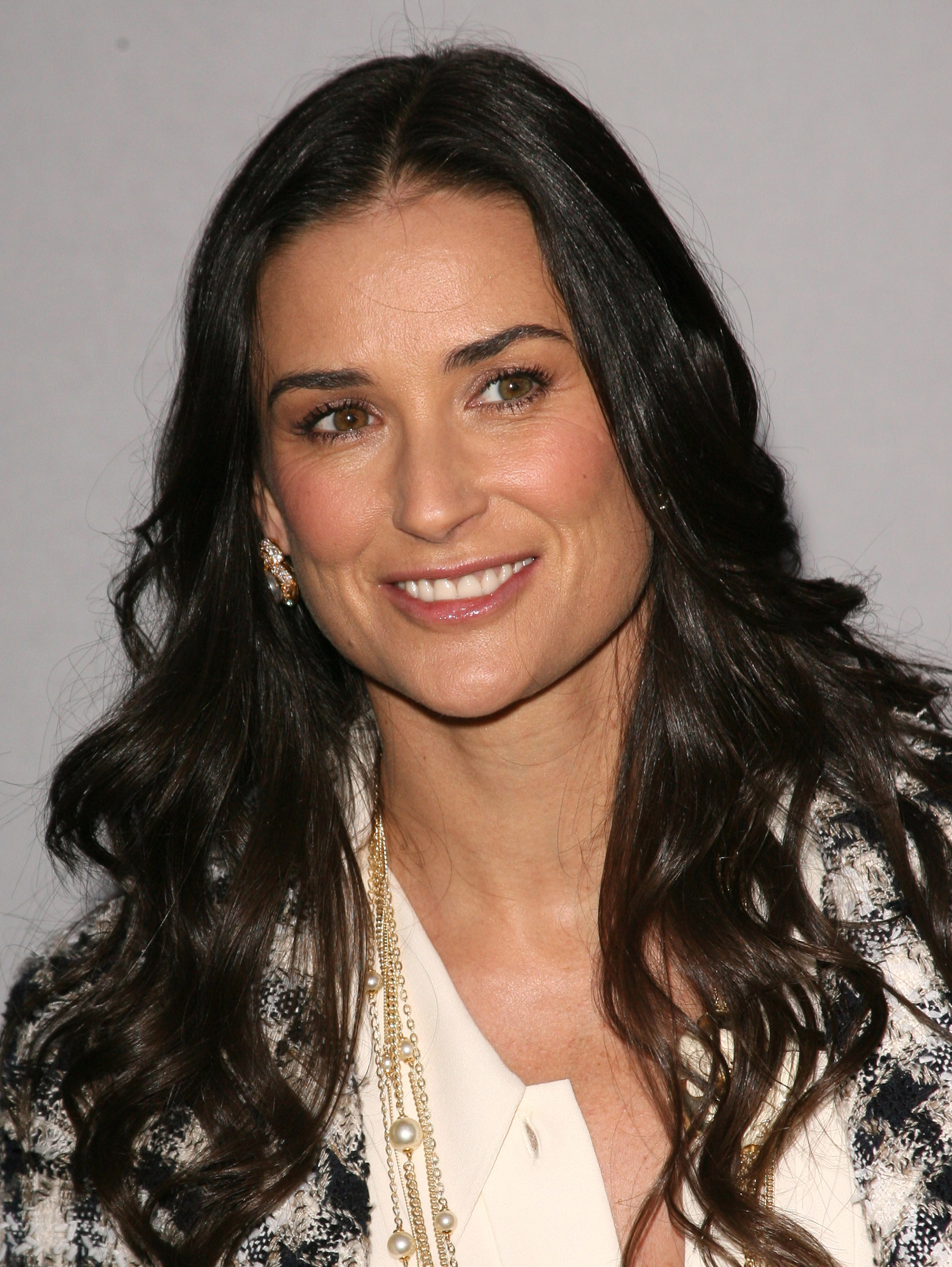 Radiating timeless grace, Demi Moore smiles with quiet confidence, her cascading dark hair framing a face that glows with poise. Draped in pearls and textured elegance, she embodies sophistication.