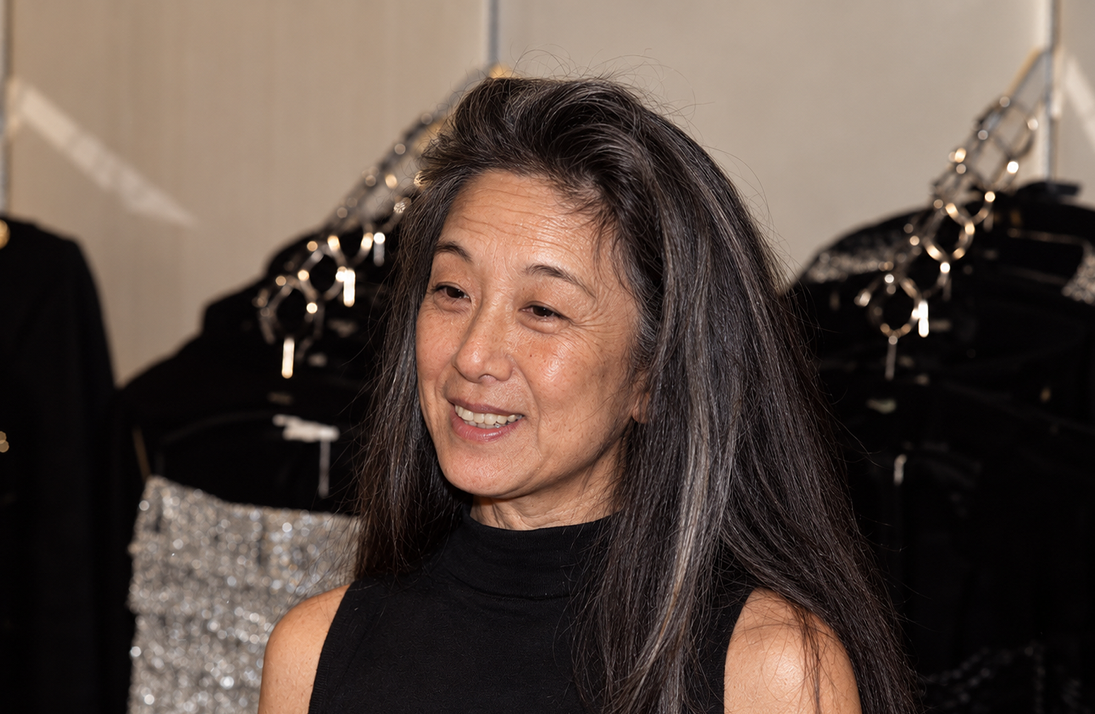 This AI creation envisions an alternate look for Vera Wang, featuring natural silver strands blended into her long dark hair, visible maturity around the eyes, and an unaltered nasal profile. Captured in what resembles a backstage showroom setting, the simulated image presents the fashion icon in a high-neck sleeveless black top with an easy, candid expression. | Source: ChatGPT