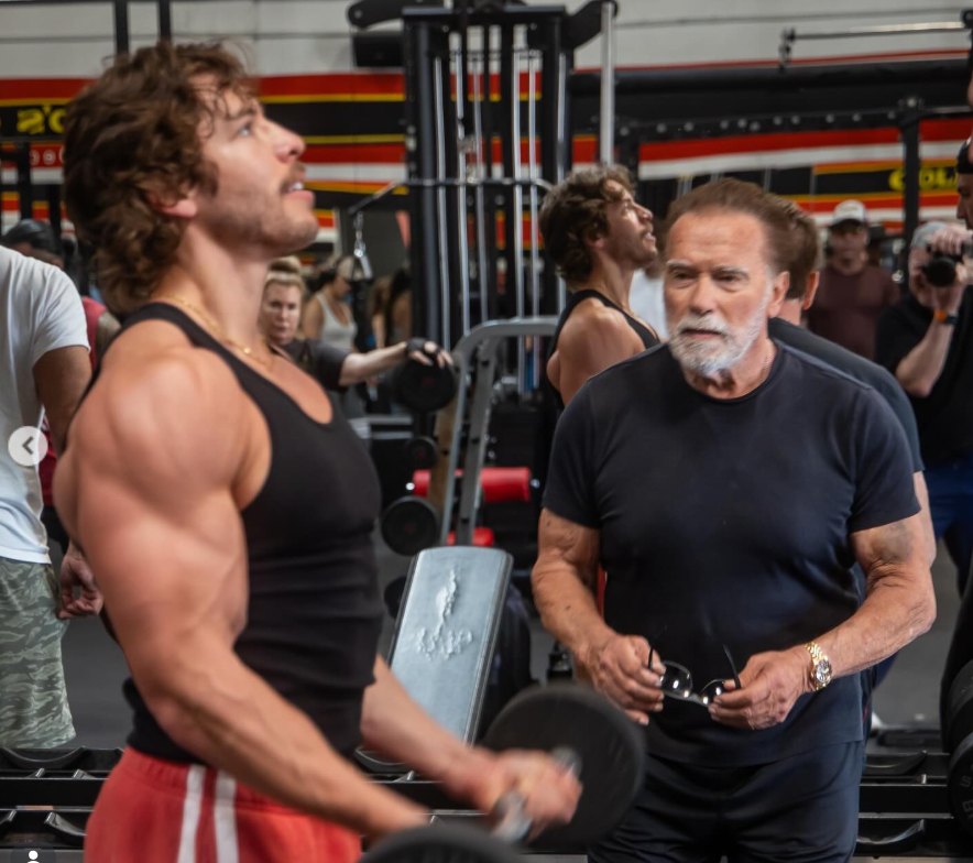 Locked in mid-rep, Joseph Baena powers through a curl as Arnold Schwarzenegger watches closely, his focused gaze capturing a moment of legacy, guidance, and shared discipline in the gym. | Source: Instagram/joebaena