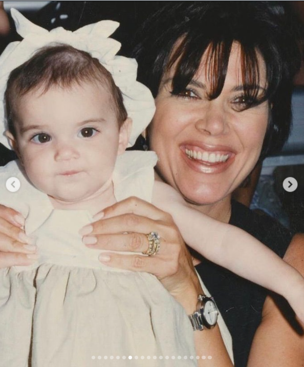 A young Kris Jenner holds baby Kendall Jenner in her arms, with the pair posing closely for a family photograph. Kris wears her hair in a short, voluminous style with bangs, while Kendall wears a light-colored dress and a large white bow. | Source: Instagram/krisjenner