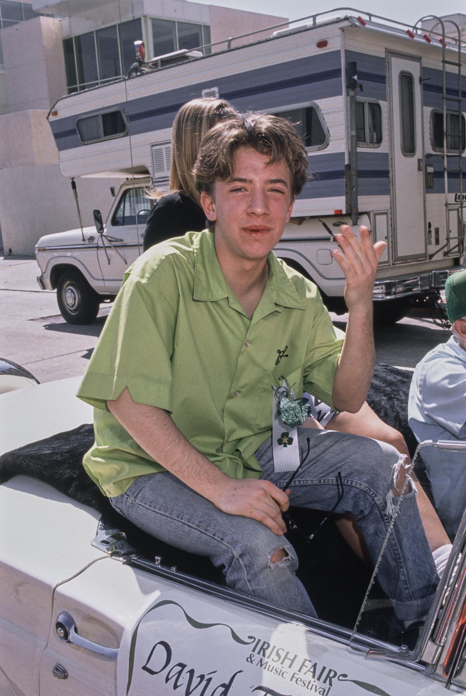 David Faustino waved to onlookers while perched on the back of an open convertible during the Irish Fair & Music Festival circa 1990 in Los Angeles, California. The "Married... with Children" star embraced a relaxed 1990s aesthetic, wearing an oversized lime-green short-sleeve button-down shirt pinned with festival ribbons and distressed, cuffed blue jeans. | Source: Getty Images
