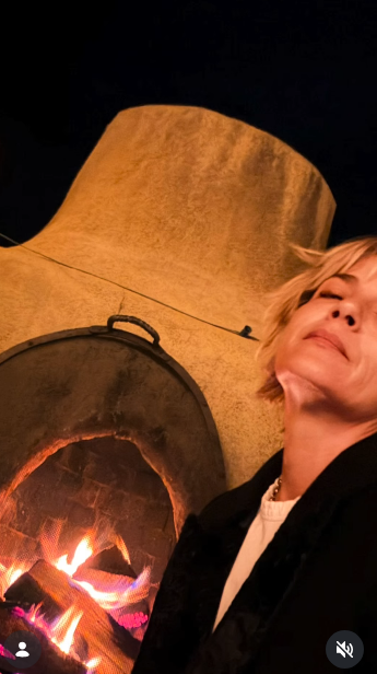 Selma Blair rests with eyes softly shut beside a lit traditional adobe kiva fireplace during a nighttime gathering on October 11, 2026. The amber firelight illuminates the rustic curved hearth as she leans back in a dark fleece coat over a white collared shirt. | Source: Instagram/selmablair