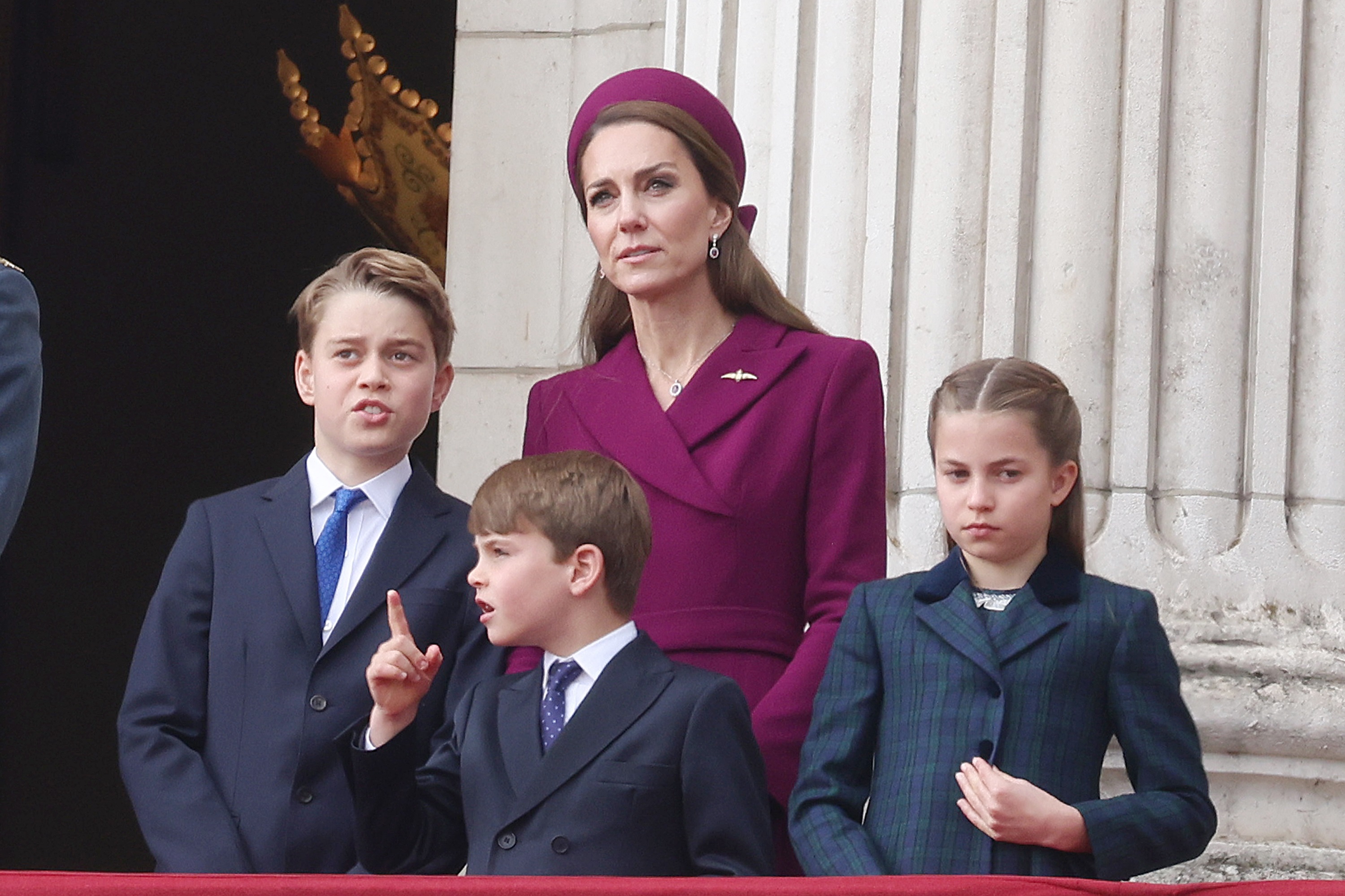 Catherine, Princess of Wales, stands on a balcony with Princess Charlotte, Prince George, and Prince Louis during the 80th anniversary of VE Day. Catherine wears a deep plum coat dress and matching hat. George and Louis are in dark suits, while Charlotte wears a dark blue plaid coat.