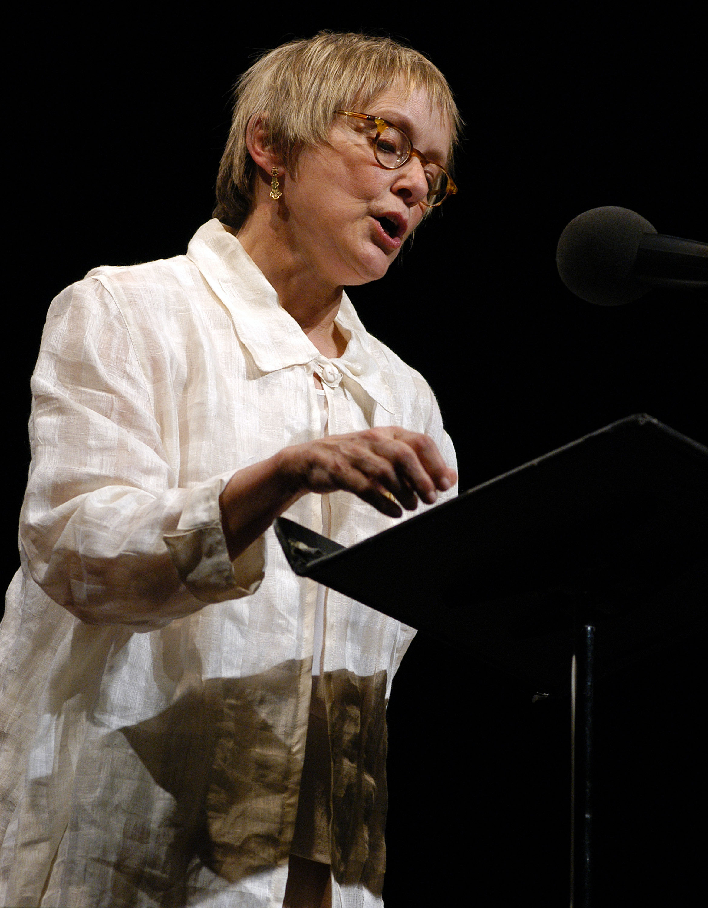 Mary Beth Hurt reads at Symphony Space in 2004 wearing a light, loose-fitting jacket and glasses. Leaning over the lectern, Hurt appears focused as she delivers a short story for the Selected Shorts event.