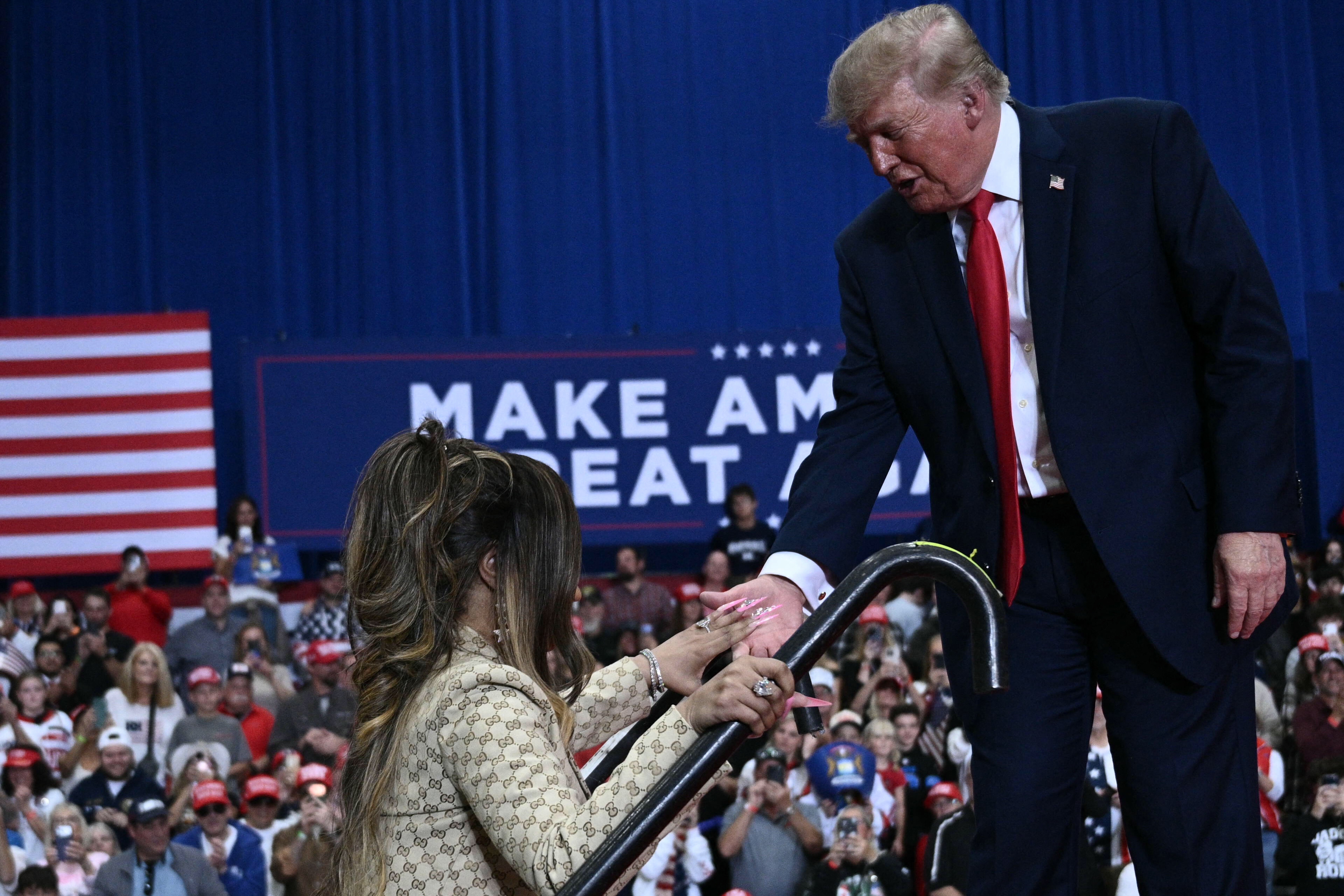 Donald Trump reaches down to help Nicki Minaj at the stage steps as she holds a microphone and steadies herself against the railing, while audience members capture the moment on their phones. | Source: Getty Images