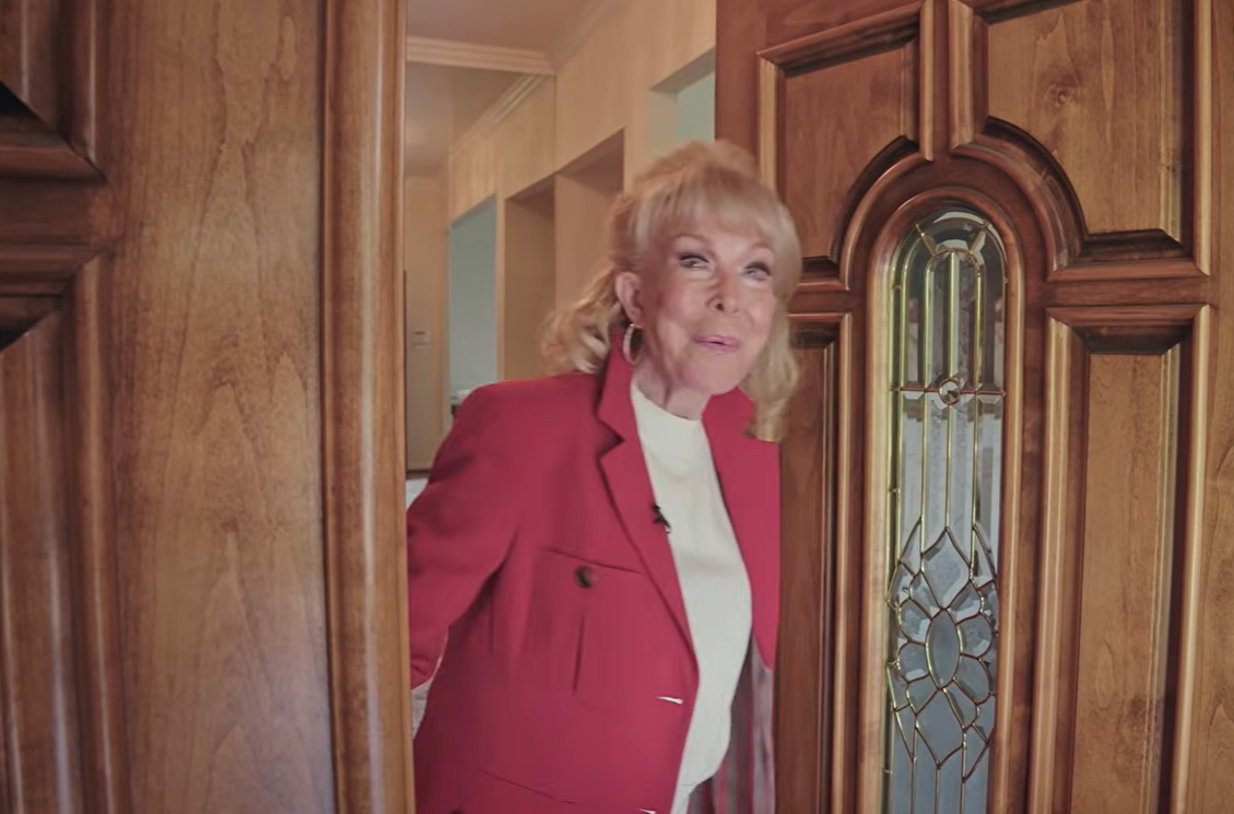 Barbara Eden smiles as she welcomes viewers into her Beverly Hills home, standing beside an ornate wooden front door with decorative stained glass while wearing a vibrant pink-red blazer over a white top. | Source: YouTube/People