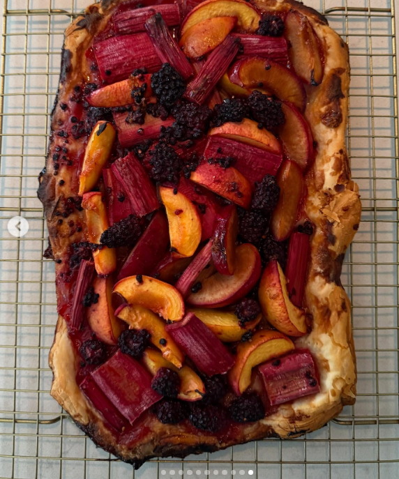A freshly baked rustic fruit tart cools on a wire rack at Arcady, crowned with roasted rhubarb, sliced stone fruits, and dark berries grown straight from the estate grounds. Reflecting Pamela Anderson's culinary passions and her plant-forward lifestyle, the artisanal bake turns seasonal garden harvests into edible art. She shared the delicious creation alongside the reflective caption, "I think of life as a garden… You get to replant, start again, and make it yours. 🤍 Love, P". | Source: Instagram/pamelaanderson