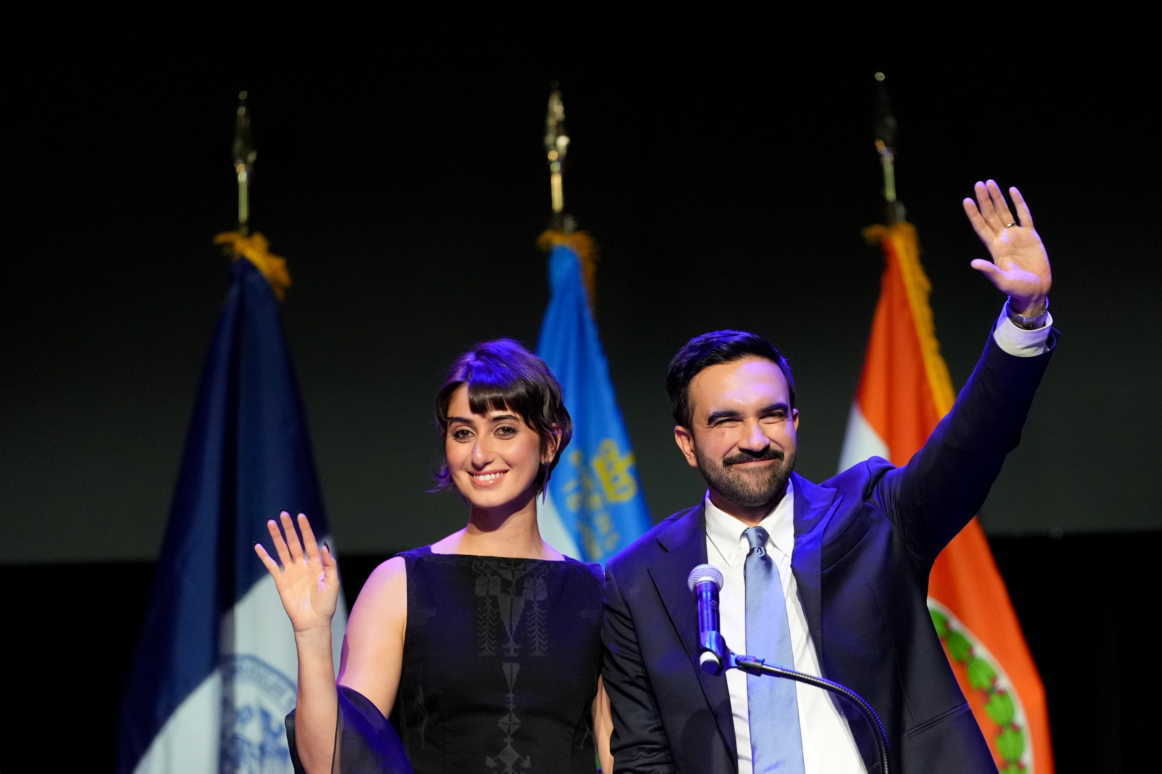 Beaming with pride, Zohra Mamdani and Rama Duwaji wave to the crowd from the stage, framed by city and state flags during a night of celebration and victory in Brooklyn.