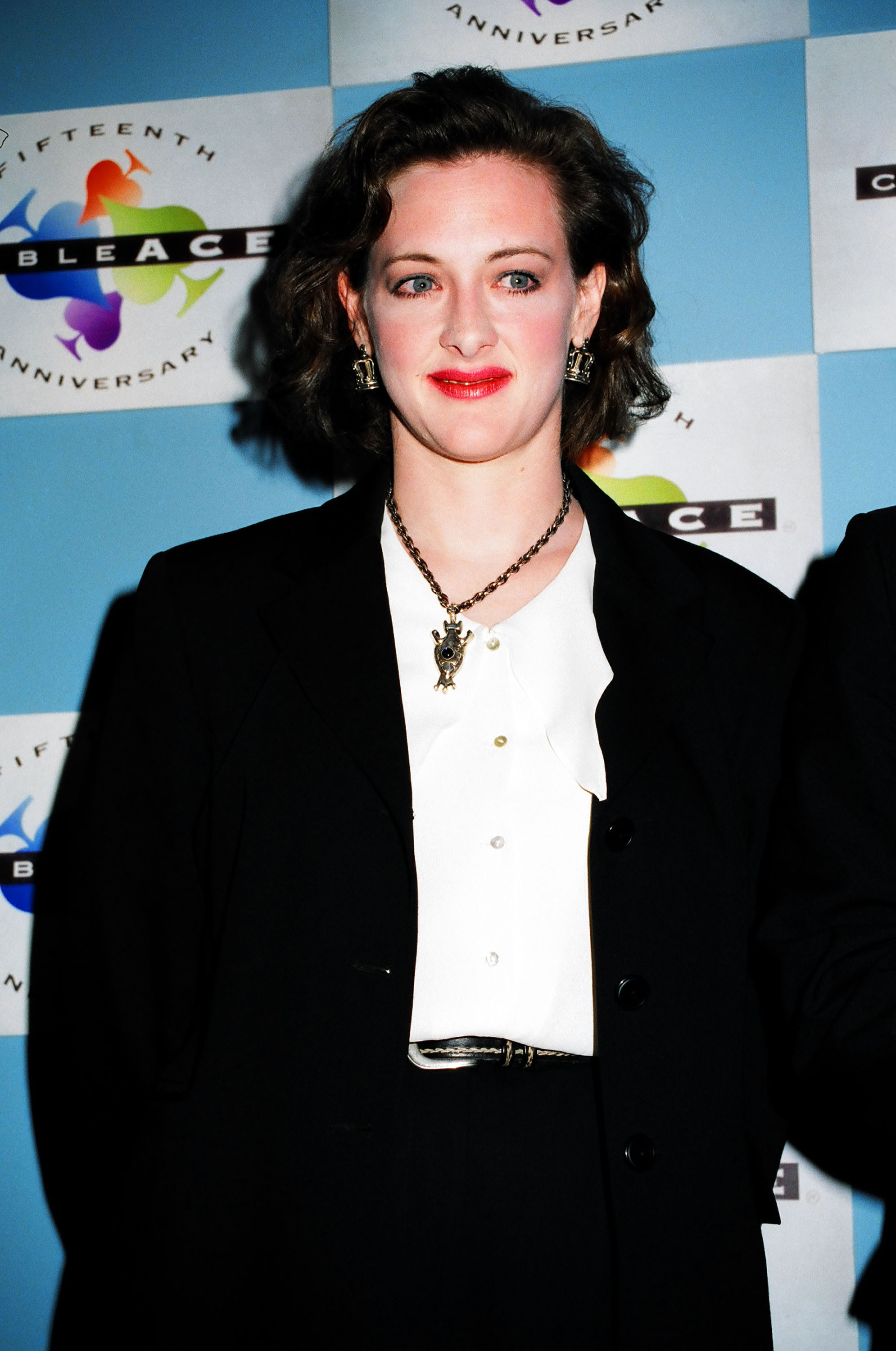 Dressed in a black blazer over a crisp white blouse, Joan Cusack strikes a polished and professional pose. A statement pendant and bold red lipstick add personality to the sophisticated look.