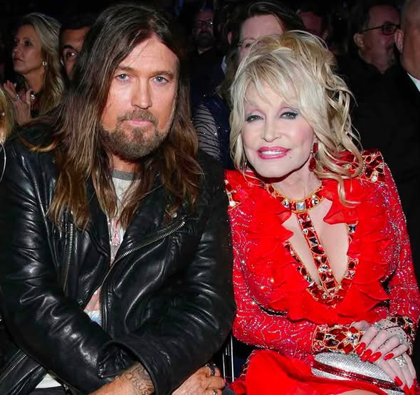 Dolly Parton, stunning in a bright red, embellished dress, sits alongside Billy Ray Cyrus, rocking his signature leather jacket and long hair, at a public event. | Source: Facebook/BillyRayCyrus