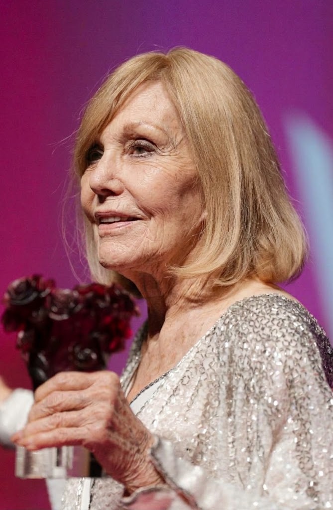 Kim Novak turns slightly in profile while holding a dark red floral award, her sparkling silver jacket catching the light against a vivid pink backdrop. | Source: Gemini
