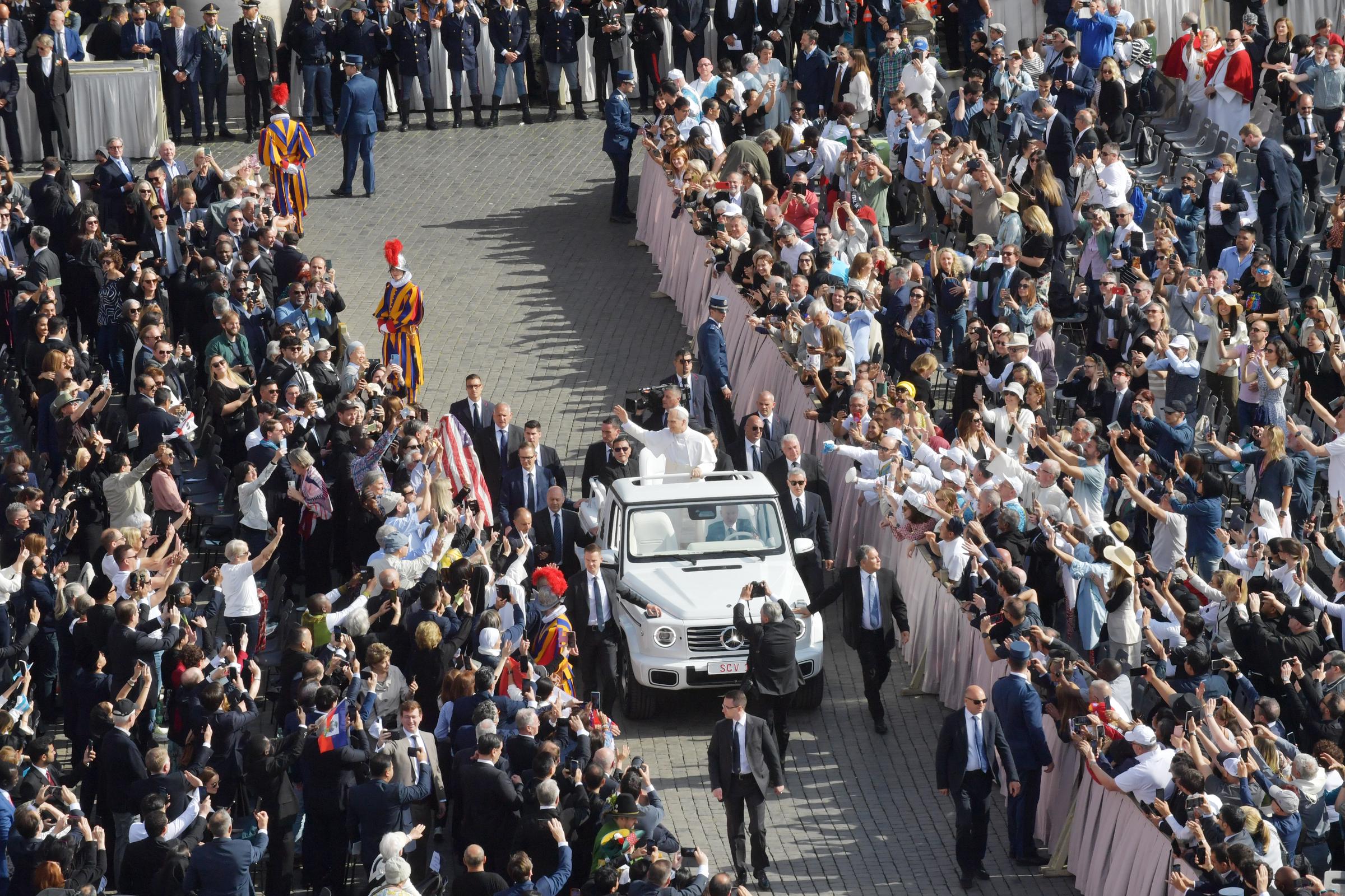 Pope Leo XIV greets thousands of pilgrims as he arrives in St. Peter's Square riding the Popemobile ahead of his inauguration mass on May 18, 2025, while the crowd cheers, waves flags, and captures the moment on mobile phones.