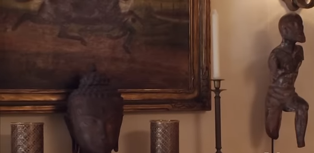 Close-up details of the fireplace mantel reveal decorative candles, a Buddha head, and unique antique sculptures. | Source: YouTube/@Biography