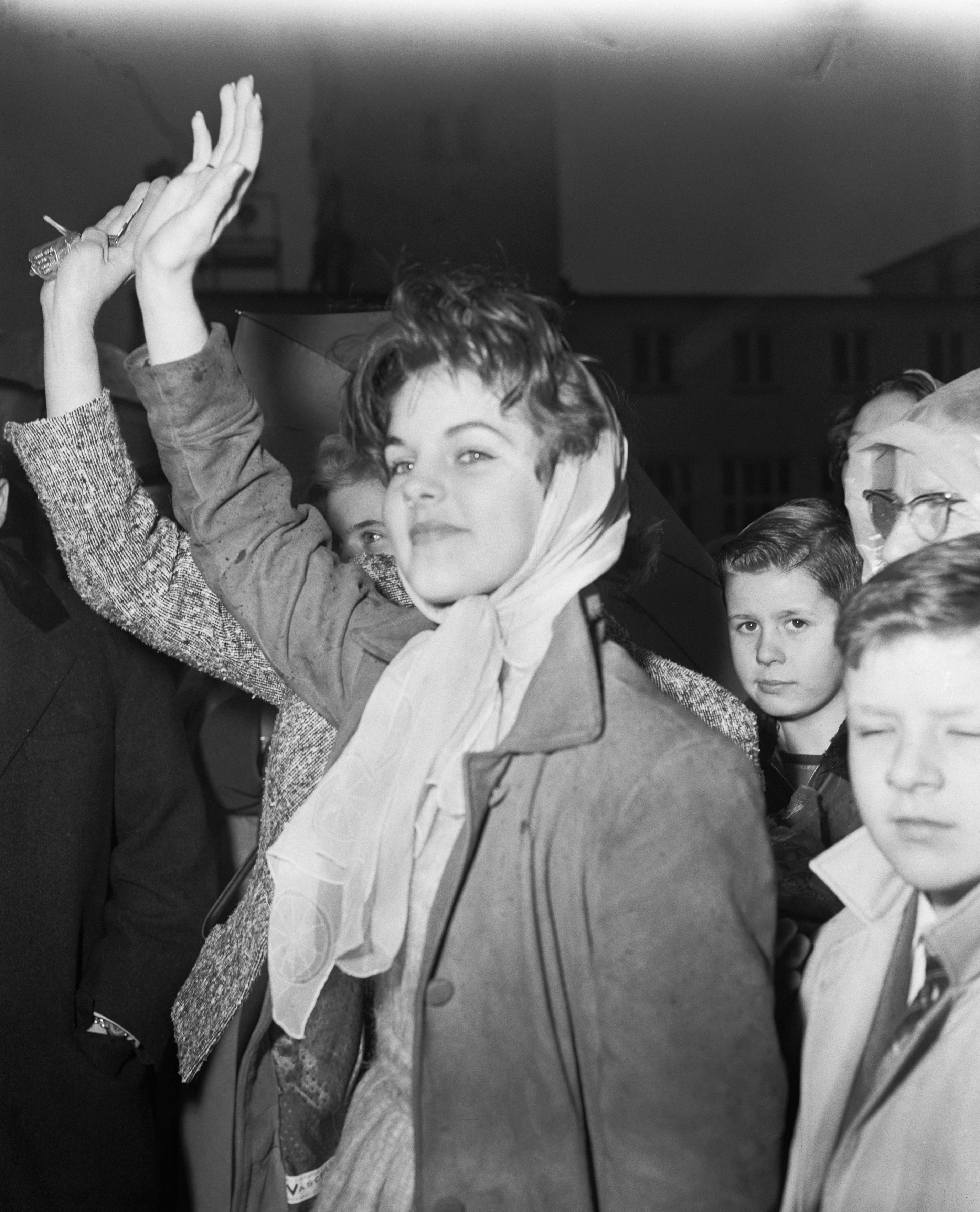 Fourteen-year-old Priscilla Beaulieu raises her hand to wave goodbye to boyfriend Elvis Presley as he departs from Rhine-Main Air Base in Frankfurt, Germany, on February 2, 1960, to return to the United States after 18 months of U.S. Army service, wearing a tailored overcoat paired with a light headscarf tied softly under her chin. | Source: Getty Images