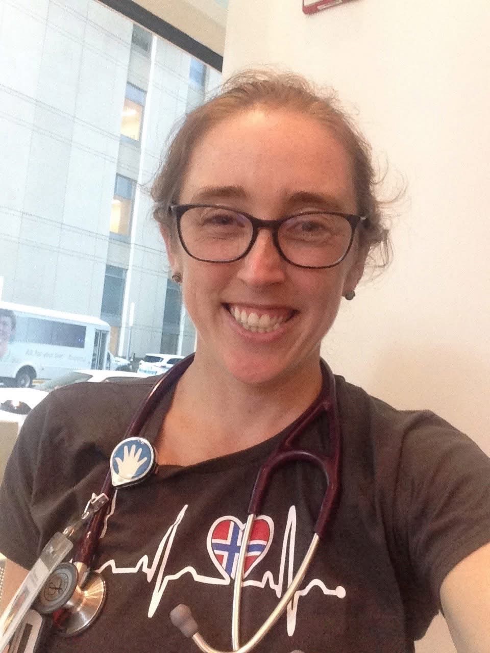 Dr. Julia Pederson smiles in a medical setting with a stethoscope draped around her neck, wearing a heartbeat graphic tee and glasses. The snapshot captures the warmth and dedication she brought to her young patients every day, standing in tragic contrast to her husband Jonathan Gatto's claims to investigators that she had stayed home unwell with a migraine on the evening of October 9, 2026, hours before emergency crews were summoned to their burning Green Street home. | Source: GoFundMe