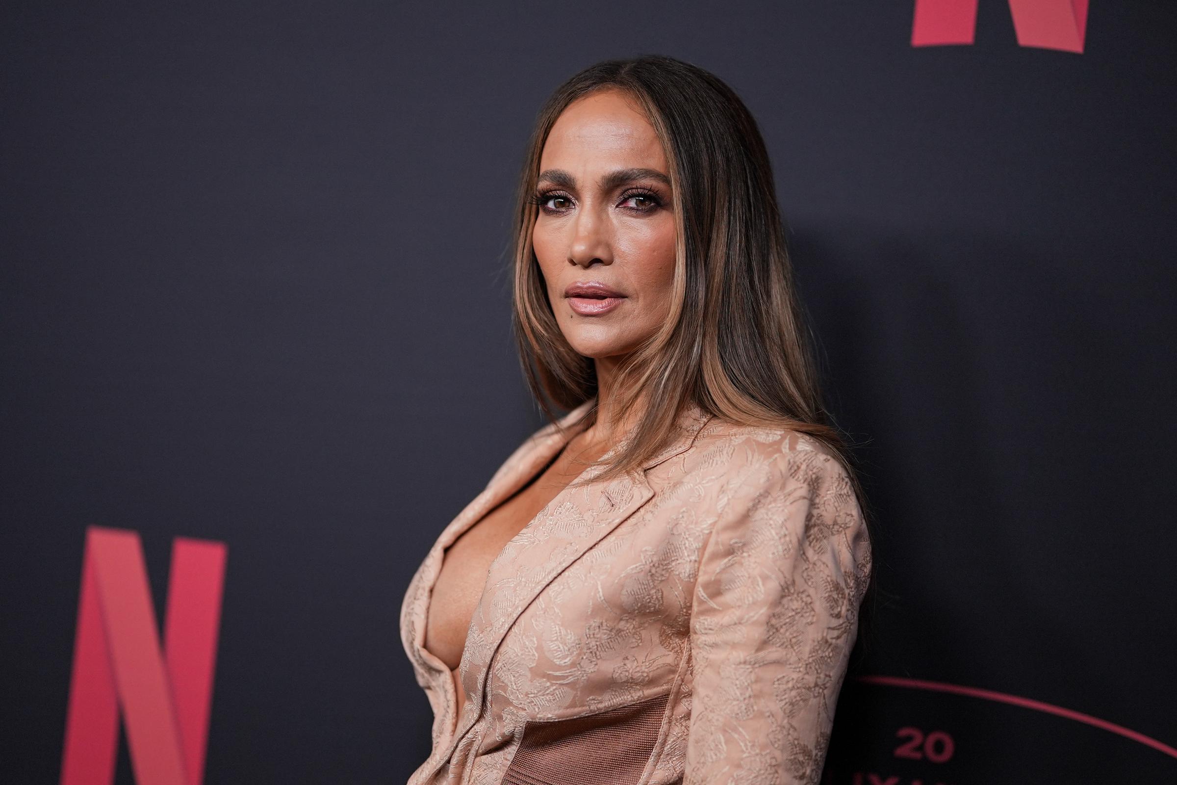 Jennifer Lopez stands angled toward the camera in a blush-toned gown featuring structured corset details, satin panels, and a plunging neckline. Her sleek straight hair, soft neutral makeup, and composed expression add elegance to the Netflix event appearance.