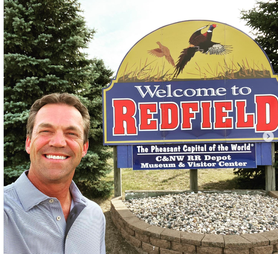 Bryon Noem smiles beside a “Welcome to Redfield” sign—marking his visit to the small town with clear pride. | Source: Instagram/sdbryonnoem