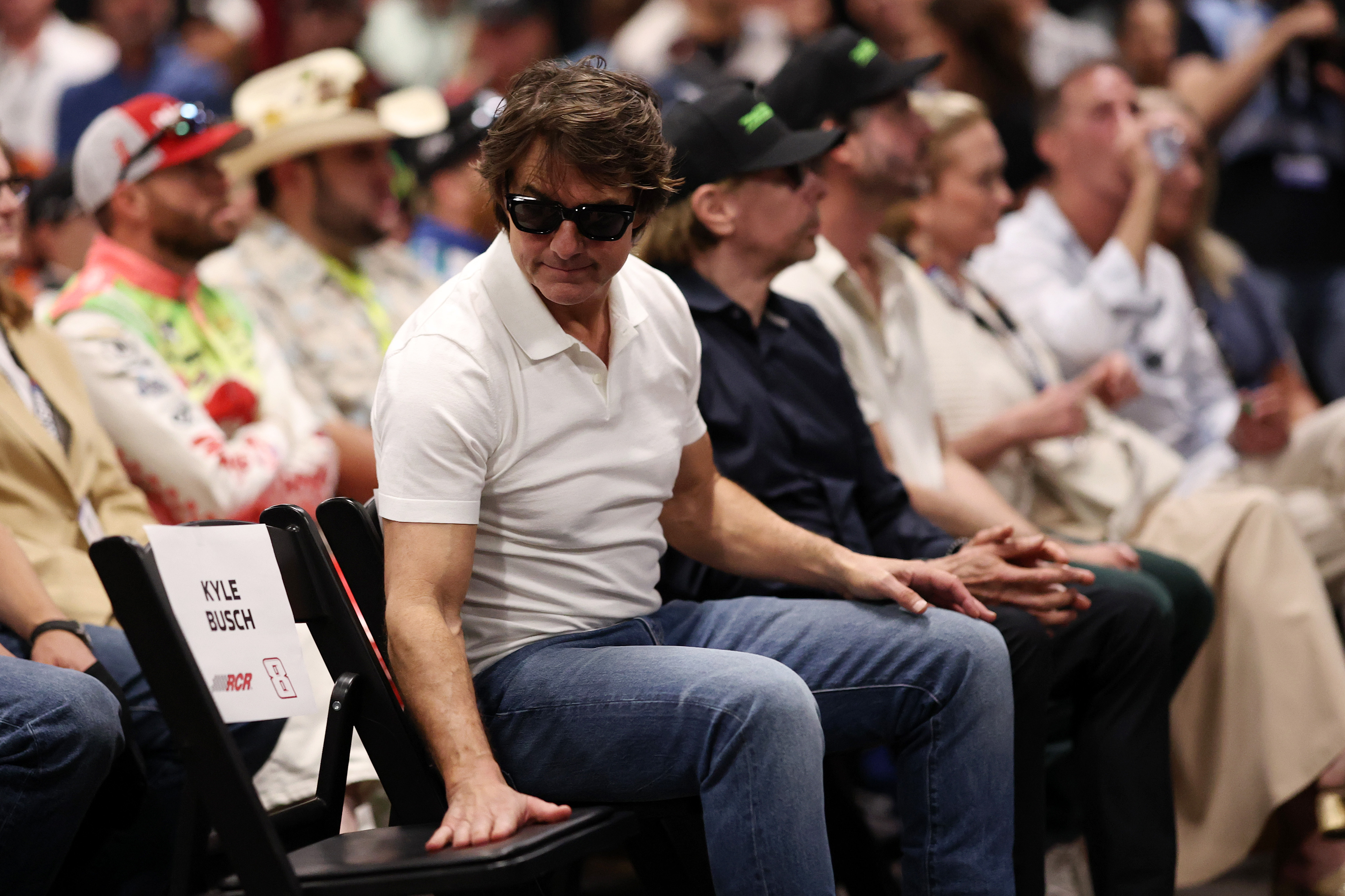 Cruise sits beside an empty chair bearing Kyle Busch's name during the drivers' meeting at Daytona International Speedway. He looks toward the seat with one hand resting on it as the gathering honors the late NASCAR champion. | Source: Getty Images
