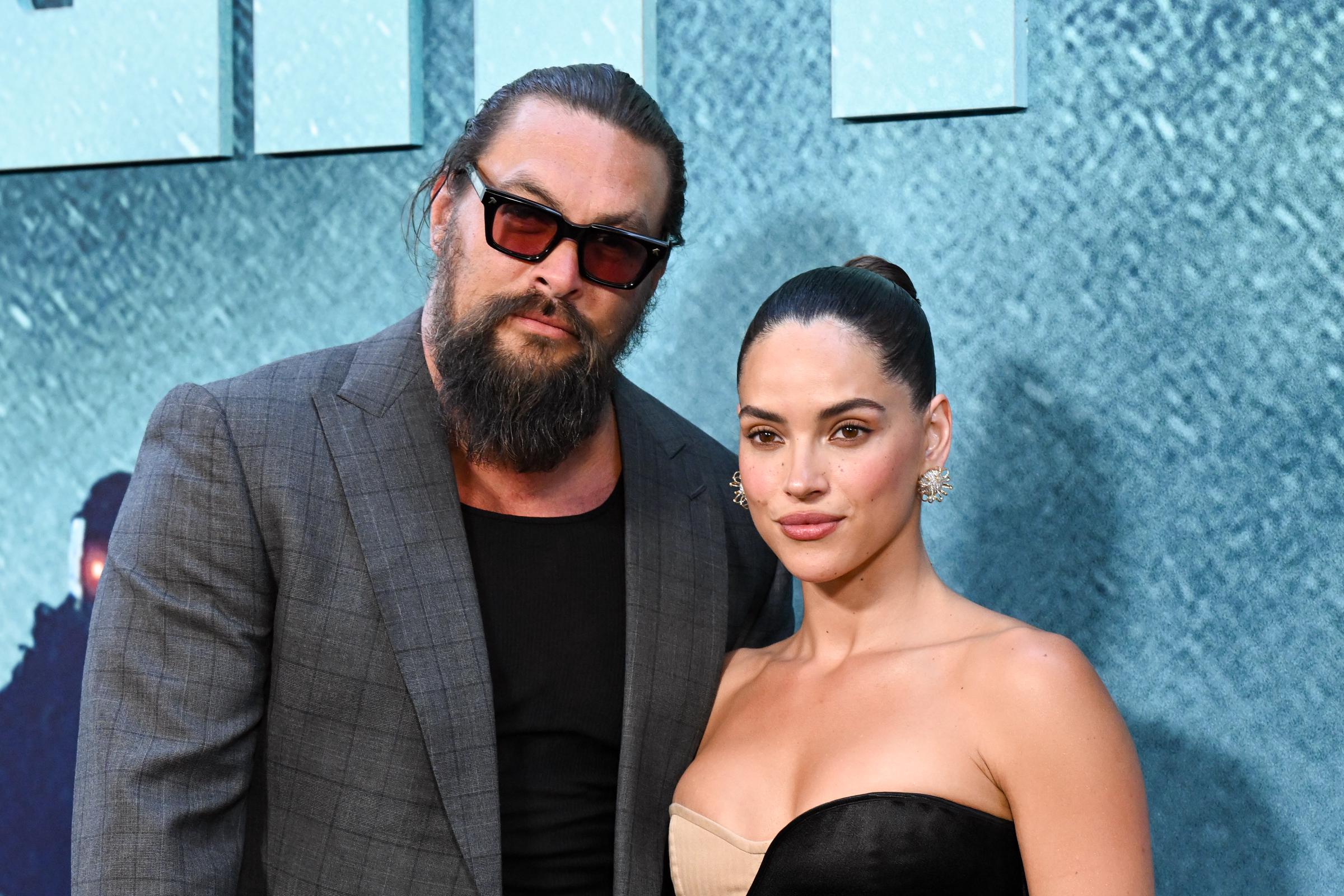 Jason Momoa and Adria Arjona attend the Los Angeles premiere of "Onslaught" at Regal L.A. Live on August 26, 2026, in Los Angeles, California. Momoa styles his hair pulled back into a sleek half-up bun with a full beard, dark-tinted sunglasses, a black scoop-neck shirt, and a grey plaid tailored suit jacket, standing close to Arjona, who wears a slicked-back high bun, ornate textured earrings, and a strapless two-tone dress featuring a structured black bodice with cream accents. | Source: Getty Images
